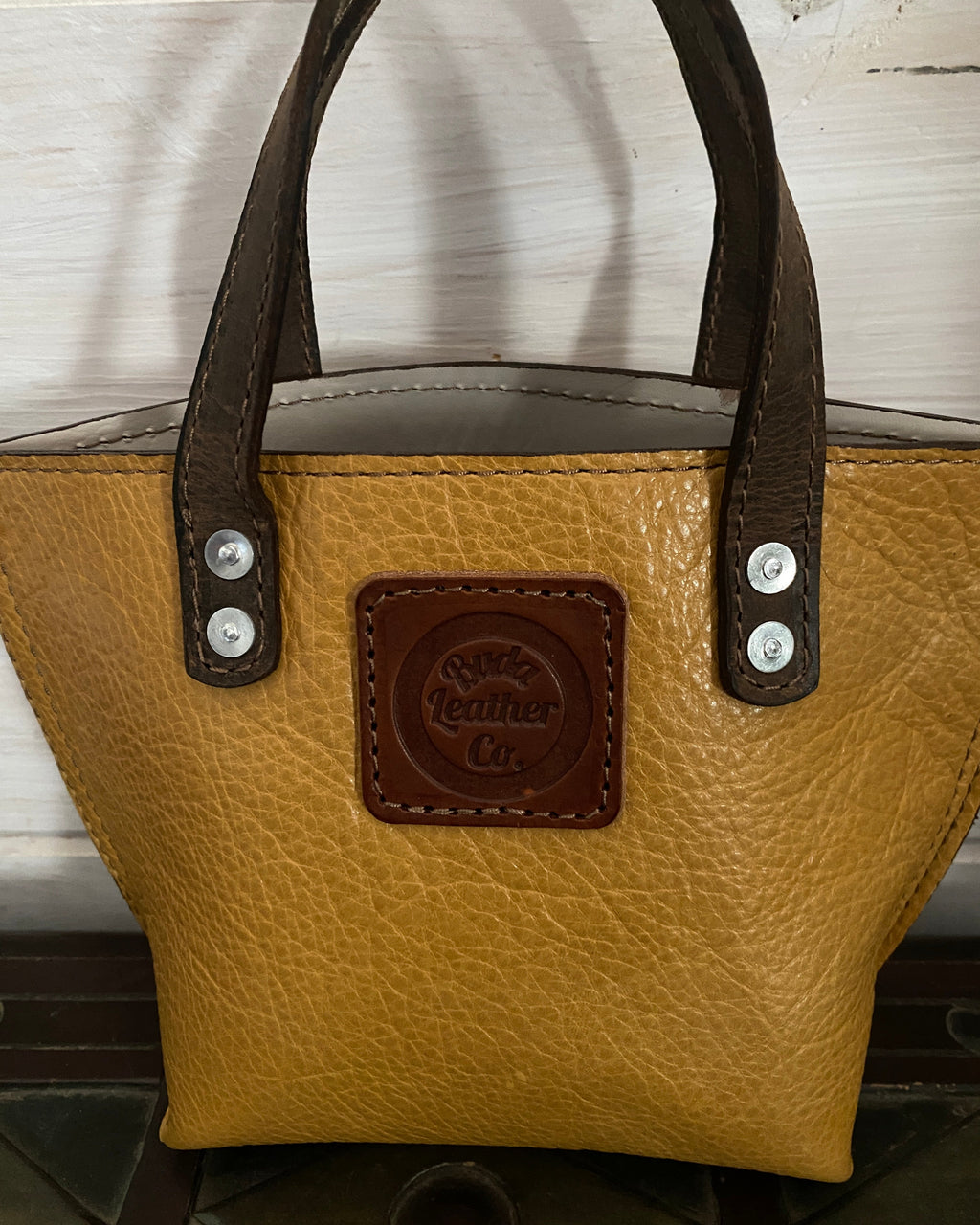 A tan pebbled leather handbag with dark brown leather handles and a matching brown embossed logo patch on the front, shown against a white wooden background.