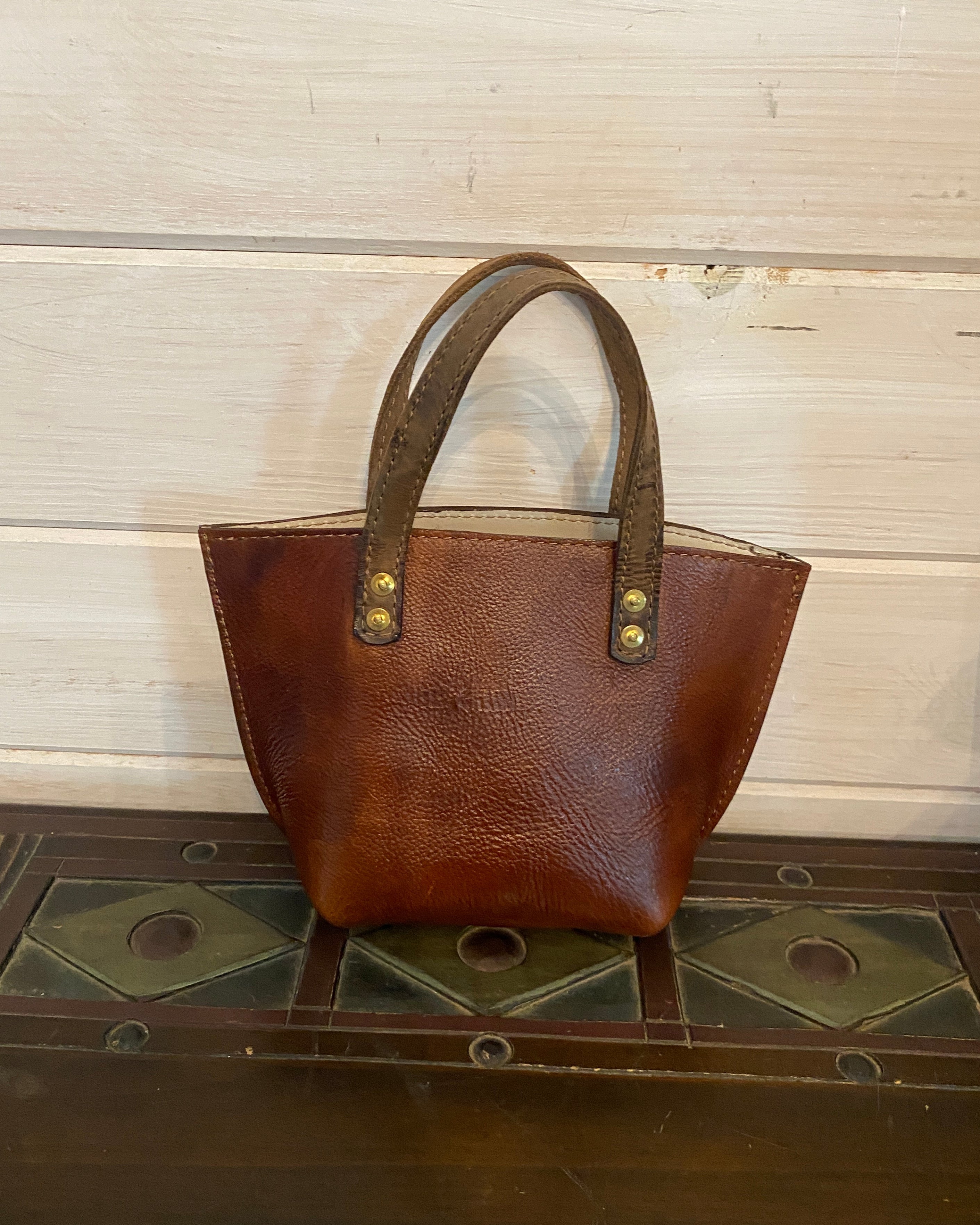 A brown leather handbag with a structured rectangular shape and two top handles, photographed against a light wood panel background next to a dark wooden surface.
