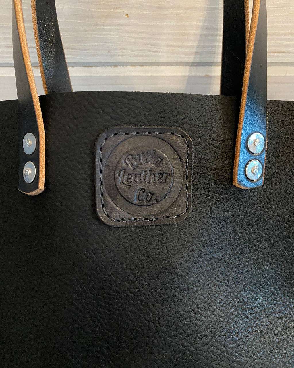 Close-up of a black leather tote bag with tan leather handles and a central embossed leather patch reading 'Buda Leather Co.'.