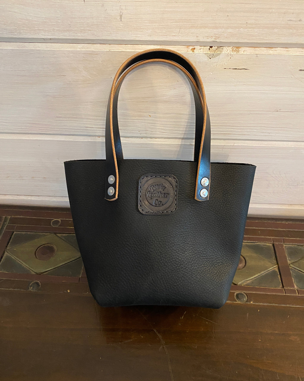 A black pebbled leather tote bag with tan leather handles and a central brand patch, displayed on a wooden surface against a light wood paneled background.