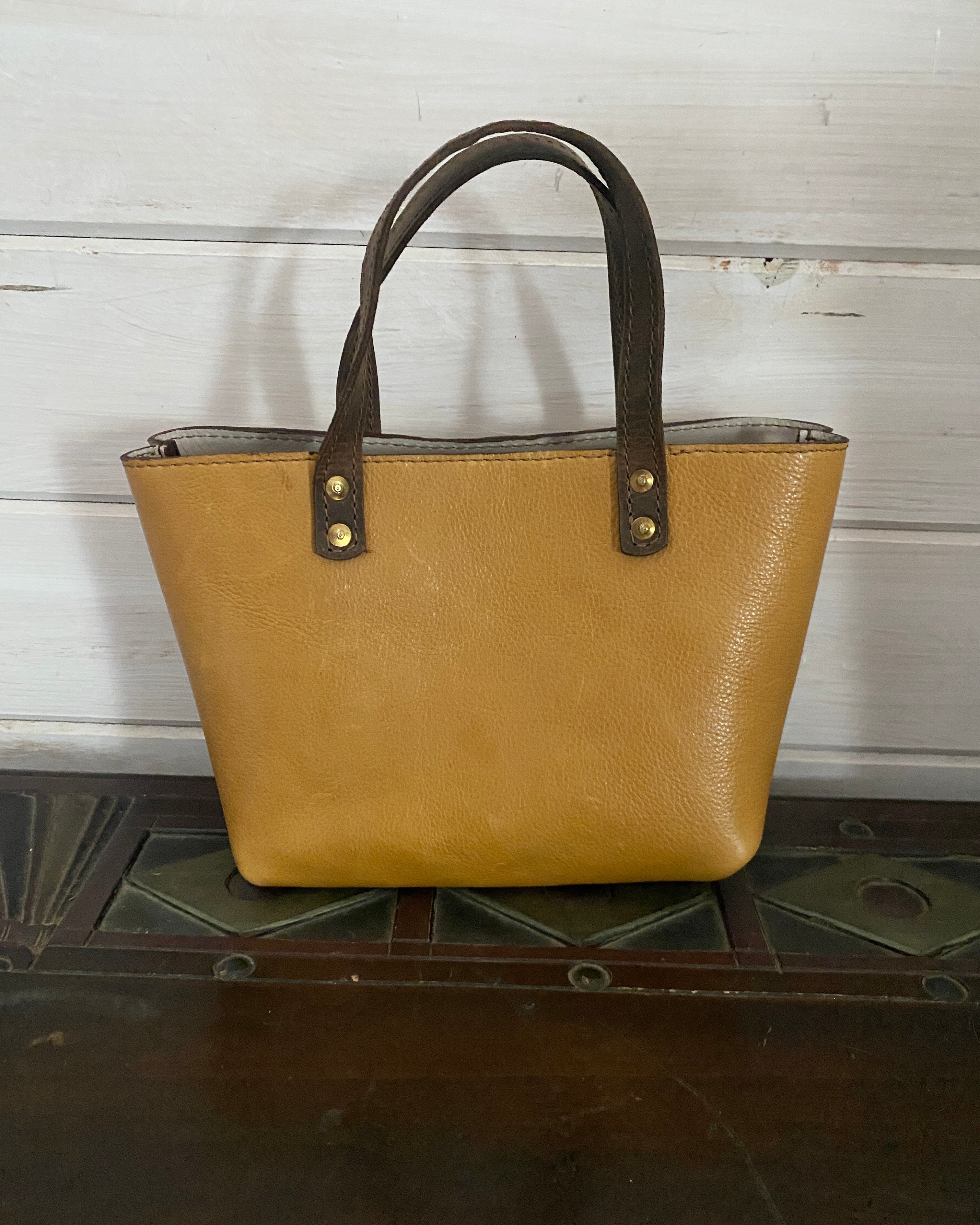 A tan pebbled leather tote bag with dark brown handles and gold-tone rivets, resting on a dark wooden surface against a white wood-paneled wall.