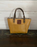 A tan pebbled leather tote bag with dark brown leather handles and a front pocket, featuring a central embossed logo patch, set against a white wood paneled background.