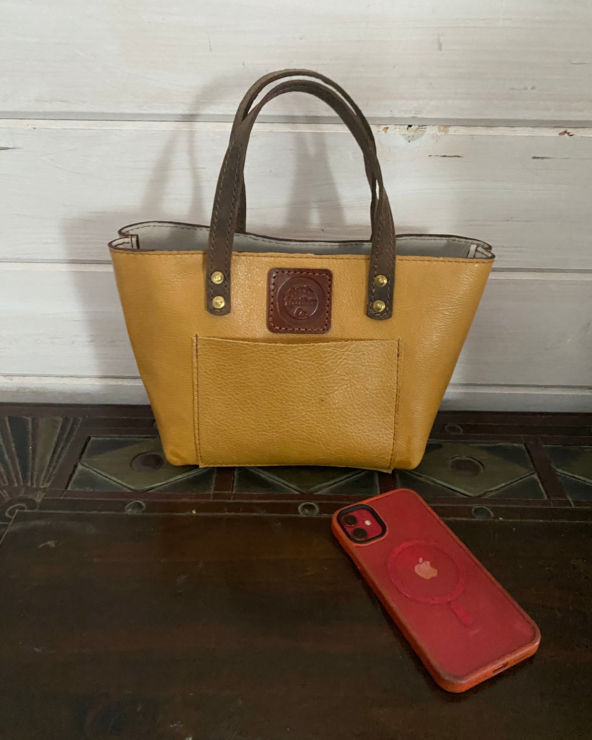 A tan leather tote bag with dark brown handles and a brown embossed logo patch on the front, displayed on a dark wooden surface next to a red phone case.