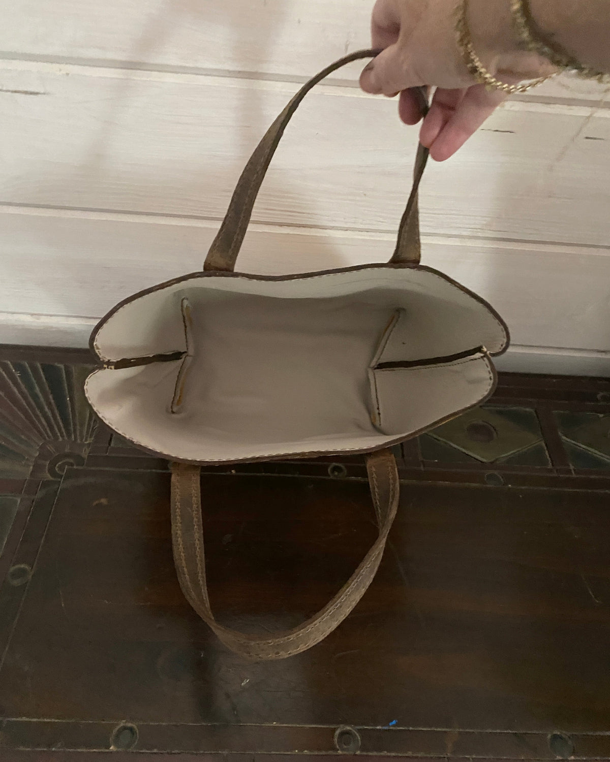 A beige tote bag with brown handles, held open by a hand with gold bracelets, set against a white paneled wall and dark wooden floor.
