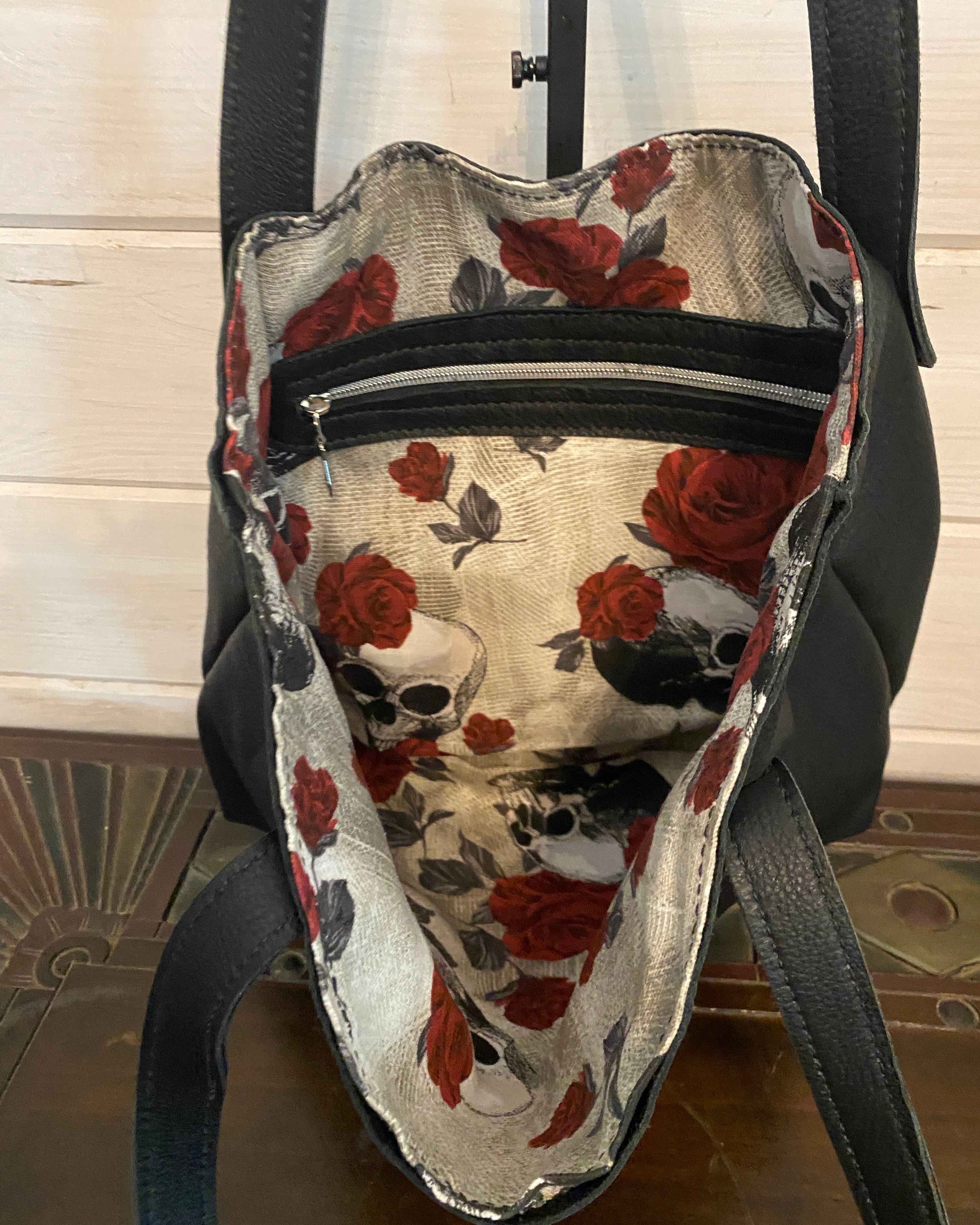 Black handbag shown from above, with the interior open to reveal a cream lining printed with red roses and skull motifs. A zipper runs vertically inside the bag, and black handles are visible against a light wood and dark wood background.