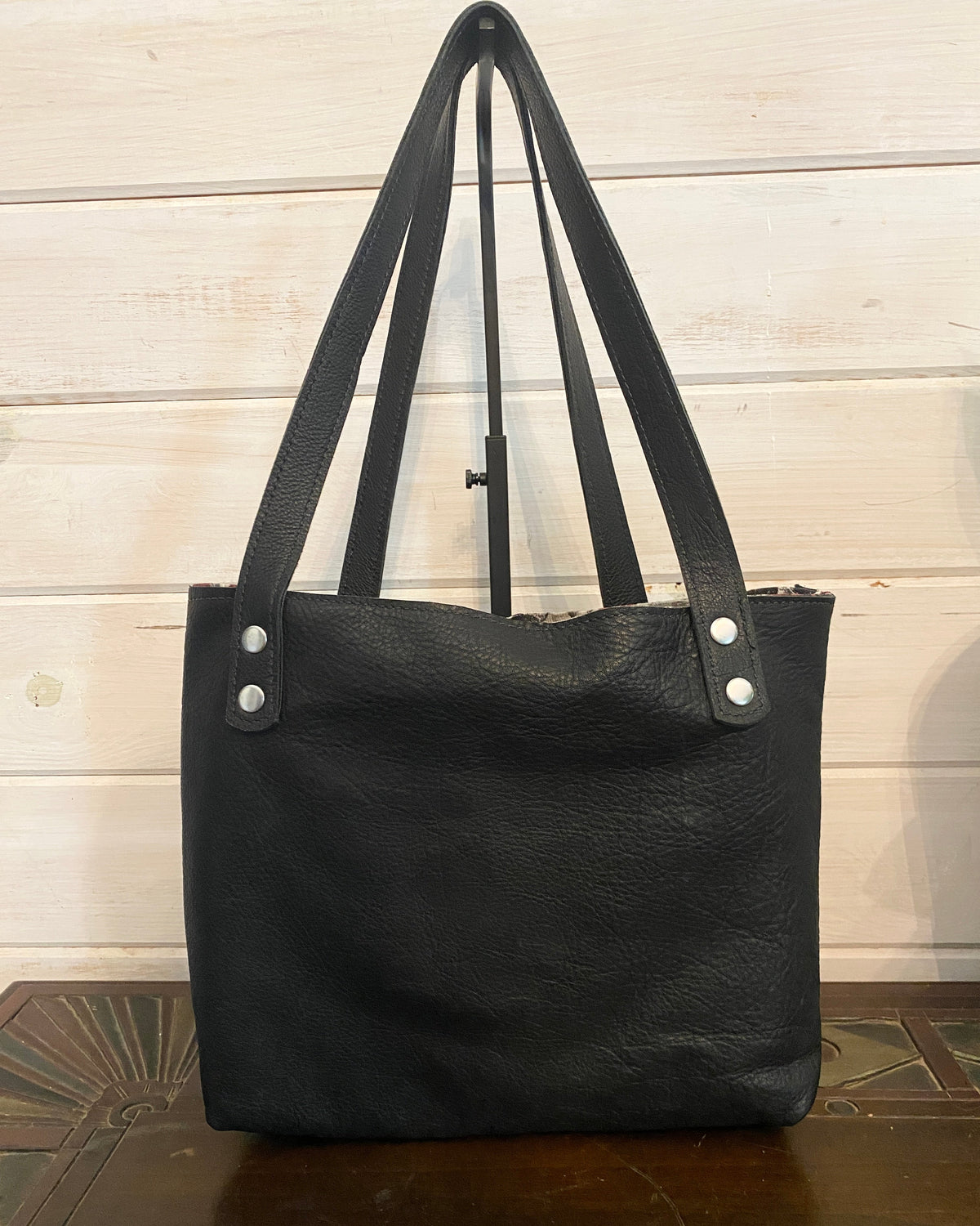 A black pebbled leather tote bag with double shoulder straps and silver-tone rivets, displayed on a wooden surface against a light wood paneled wall.