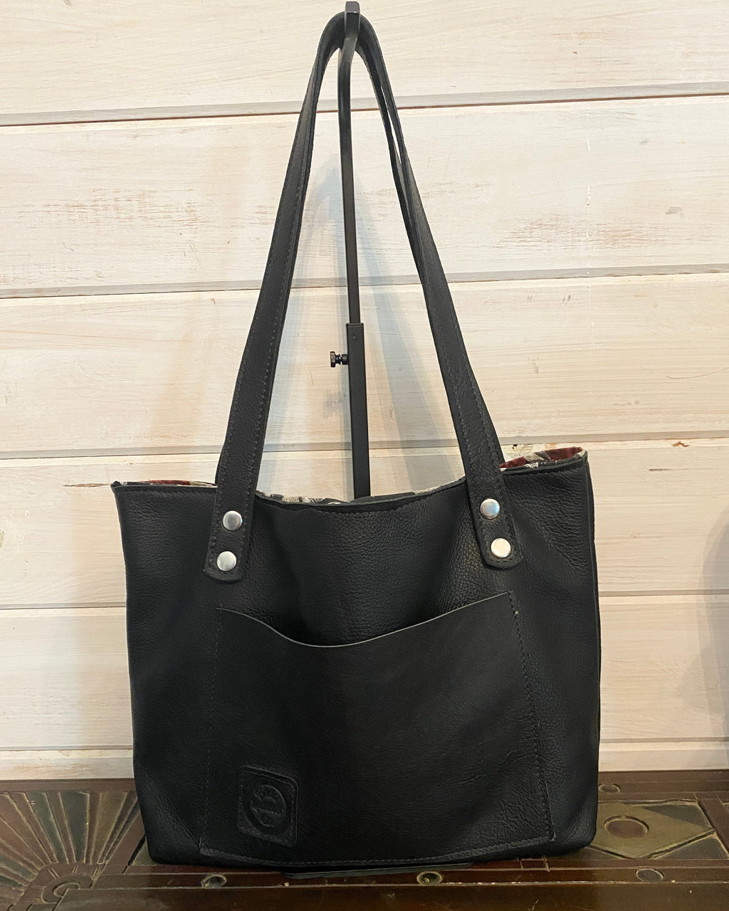 A black leather tote bag with dual shoulder straps and silver-tone snap hardware, displayed on a wooden stand against a light wood paneled wall.
