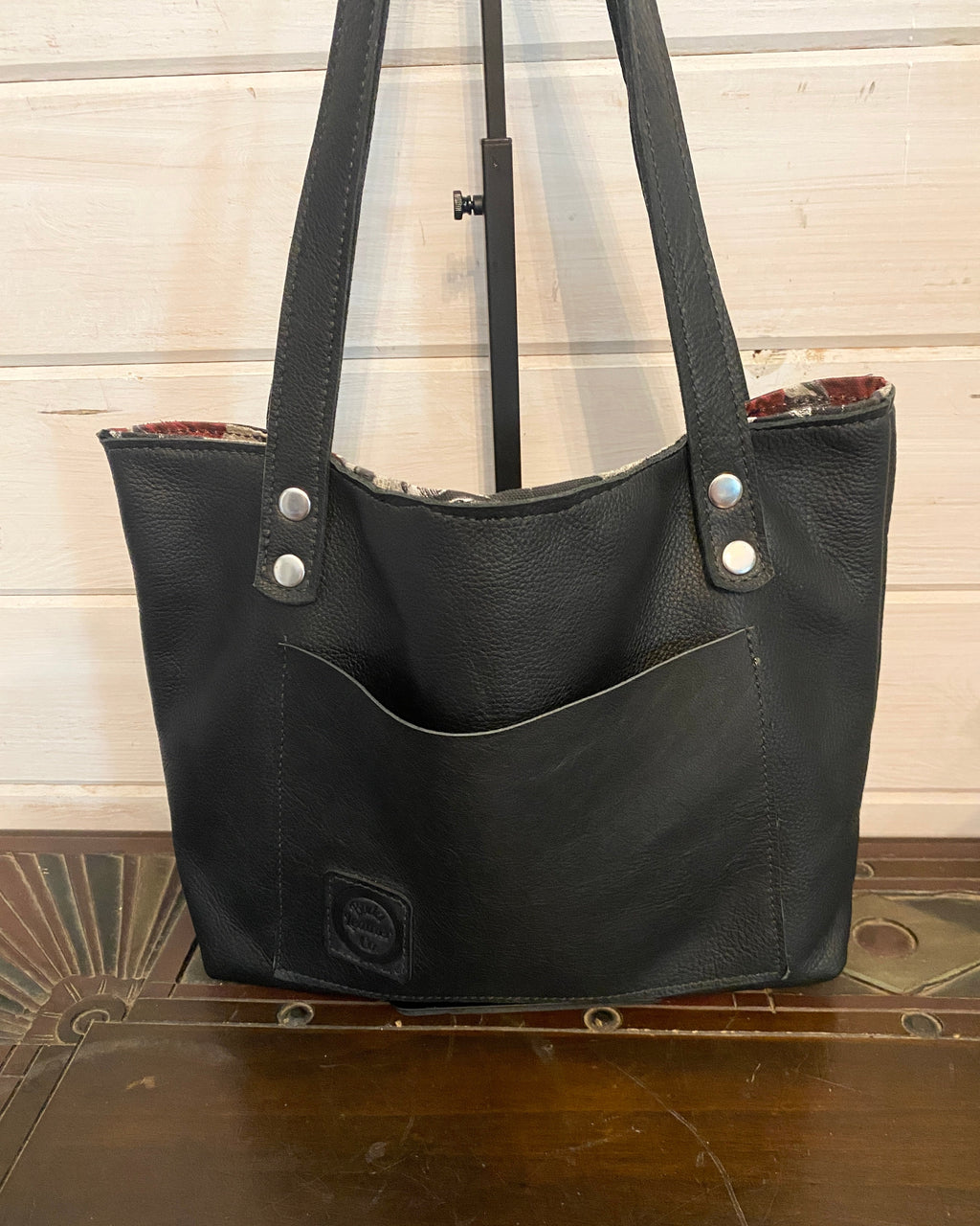 Black leather tote bag with double shoulder straps, silver-tone rivets, and a front slip pocket, displayed on a wooden surface against a light wood paneled wall.