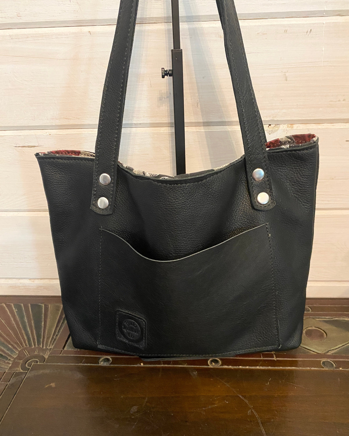 Black leather tote bag with double shoulder straps, silver-tone rivets, and a front slip pocket, displayed on a wooden surface against a light wood paneled wall.