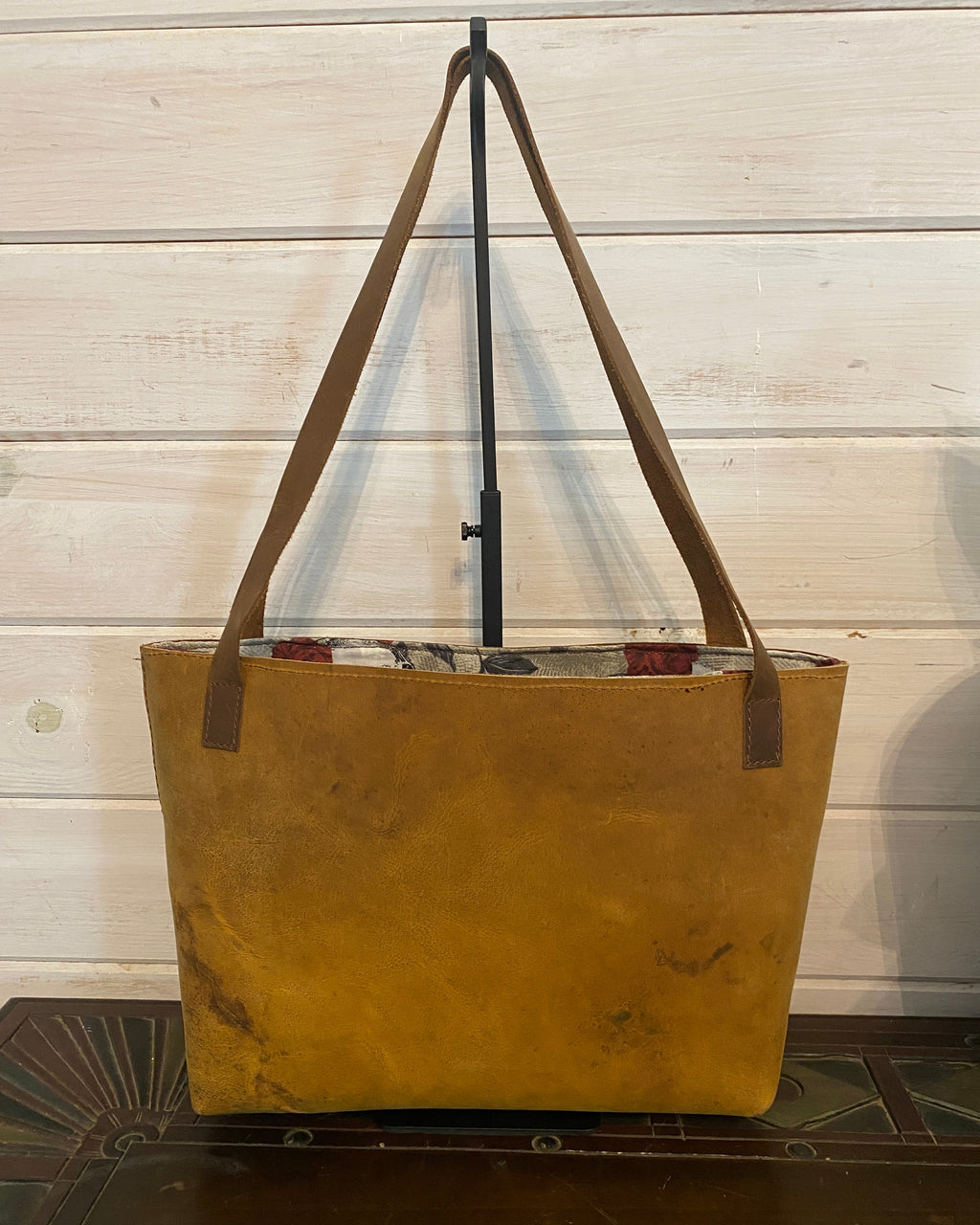A large tan canvas tote bag with brown shoulder straps, shown hanging from a black stand against a white wooden paneled wall.