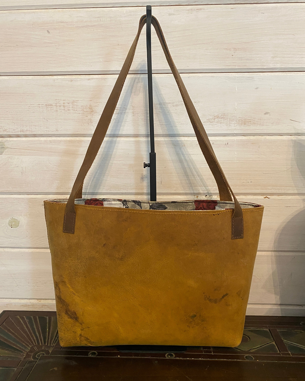 A large tan canvas tote bag with brown shoulder straps, shown hanging from a black stand against a white wooden paneled wall.