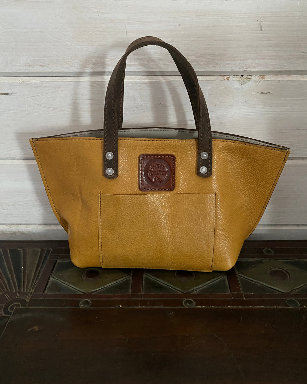 A mustard yellow leather tote bag with dark brown handles and a brown logo patch on the front pocket, displayed on a dark wooden surface against a white wood-paneled wall.