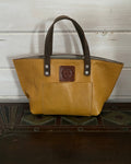 A mustard yellow leather tote bag with dark brown handles and a brown logo patch on the front pocket, displayed on a dark wooden surface against a white wood-paneled wall.