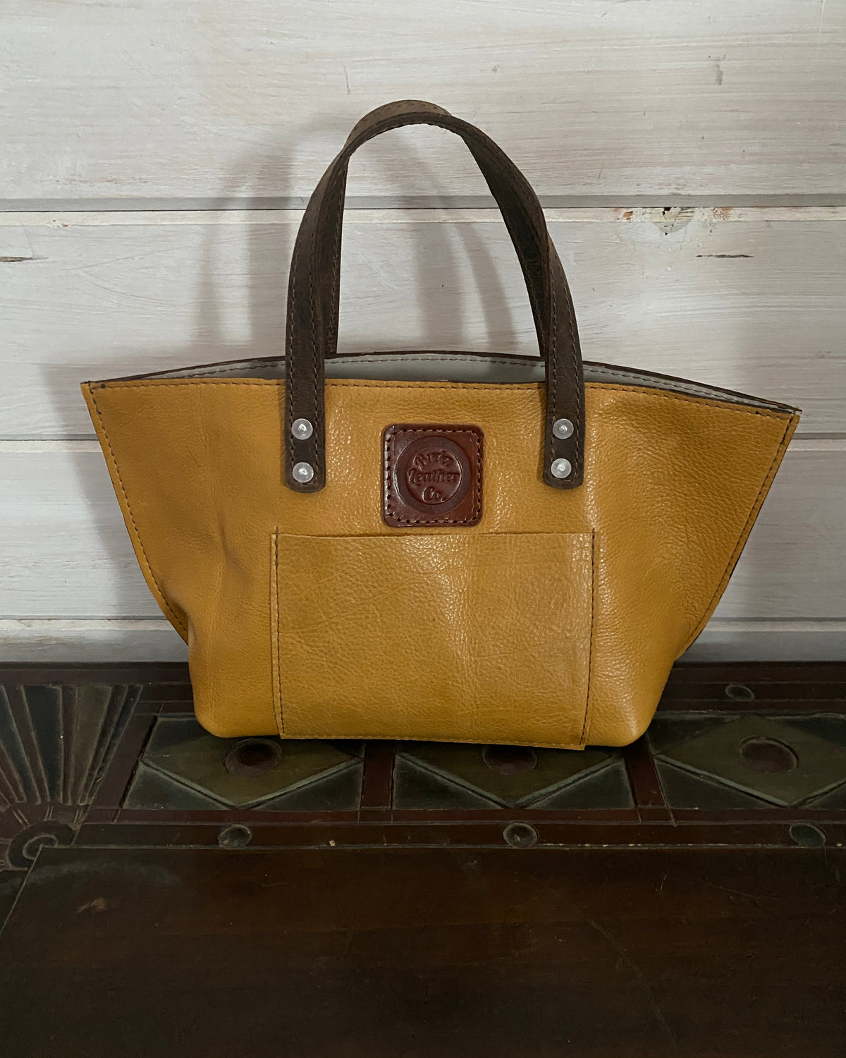 A mustard yellow leather tote bag with dark brown handles and a brown logo patch on the front pocket, displayed on a dark wooden surface against a white wood-paneled wall.