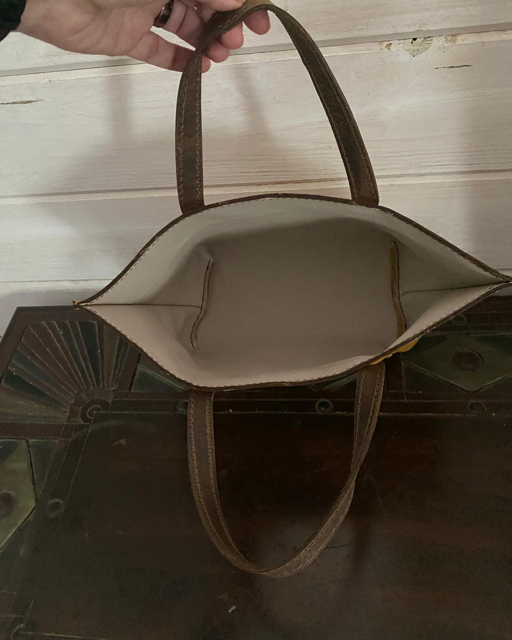 A brown leather tote bag with a cream-colored interior lining, held by a hand against a background of a white wooden door and a dark mirror.