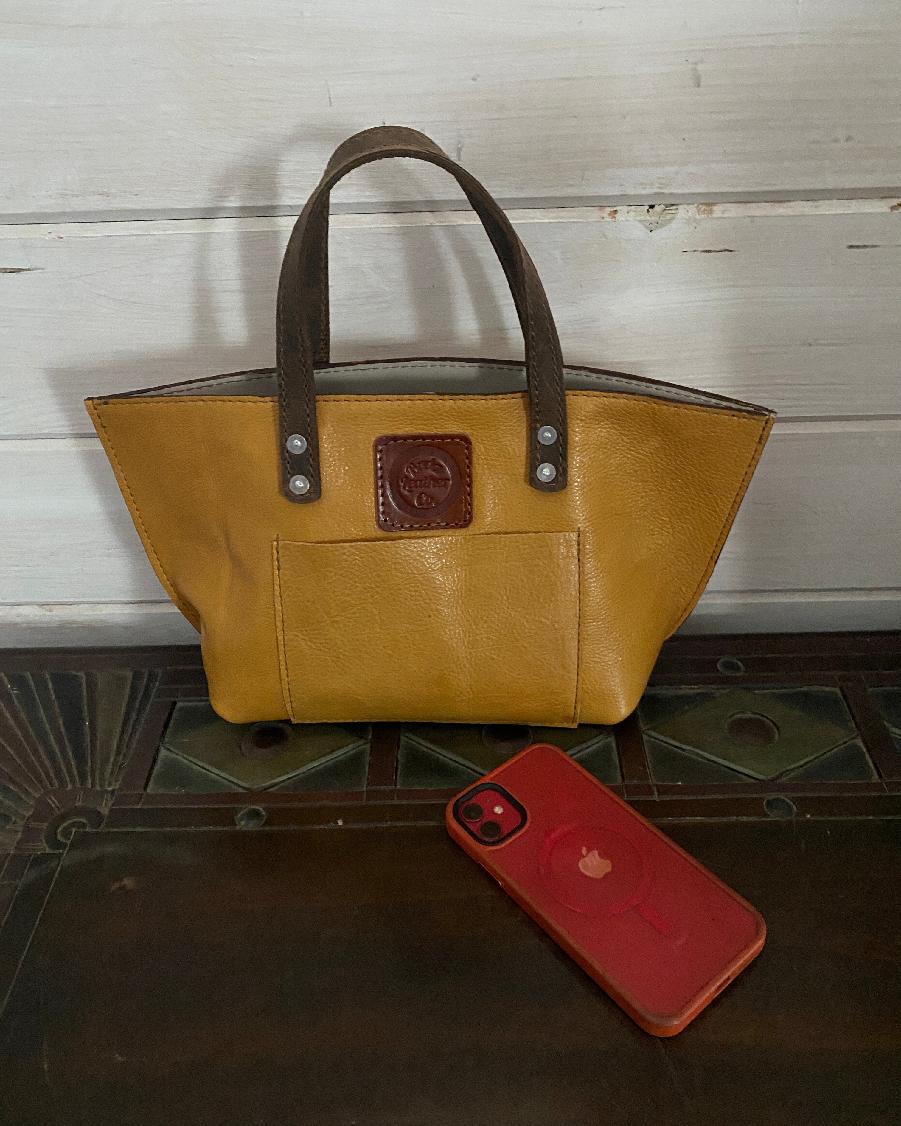 A mustard yellow leather tote bag with dark brown handles and a brown leather logo patch, displayed on a dark wooden surface next to a red smartphone case.