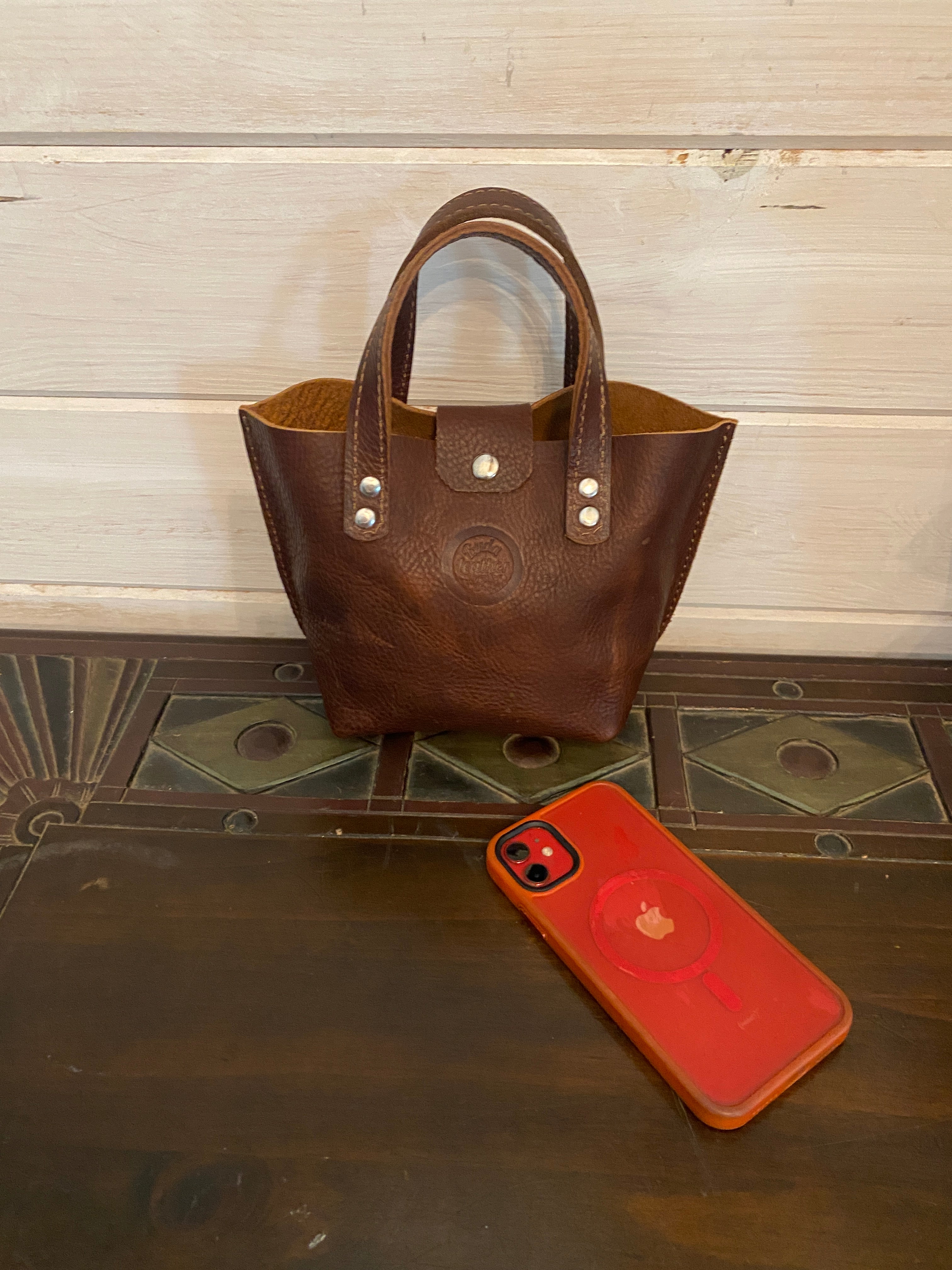 A brown leather handbag with silver-tone hardware, shown next to a red phone case on a dark wooden surface.