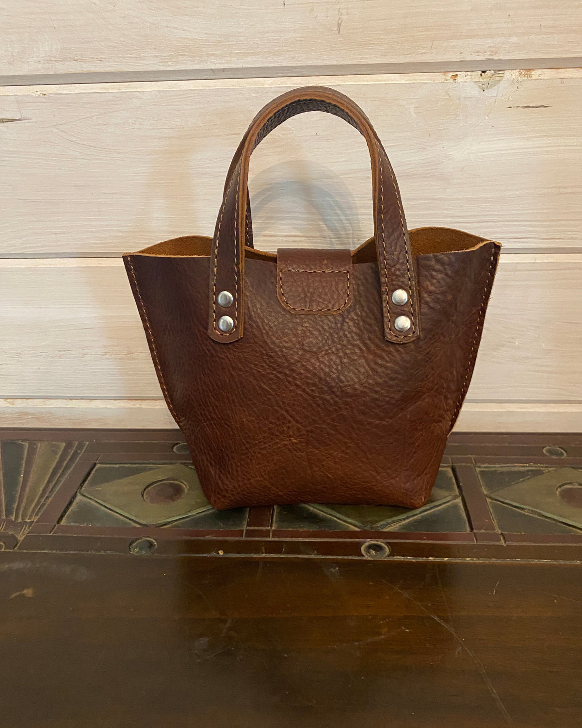 A small brown leather handbag with a pebbled texture and double top handles, shown standing on a dark wooden surface against a light wood paneled background.