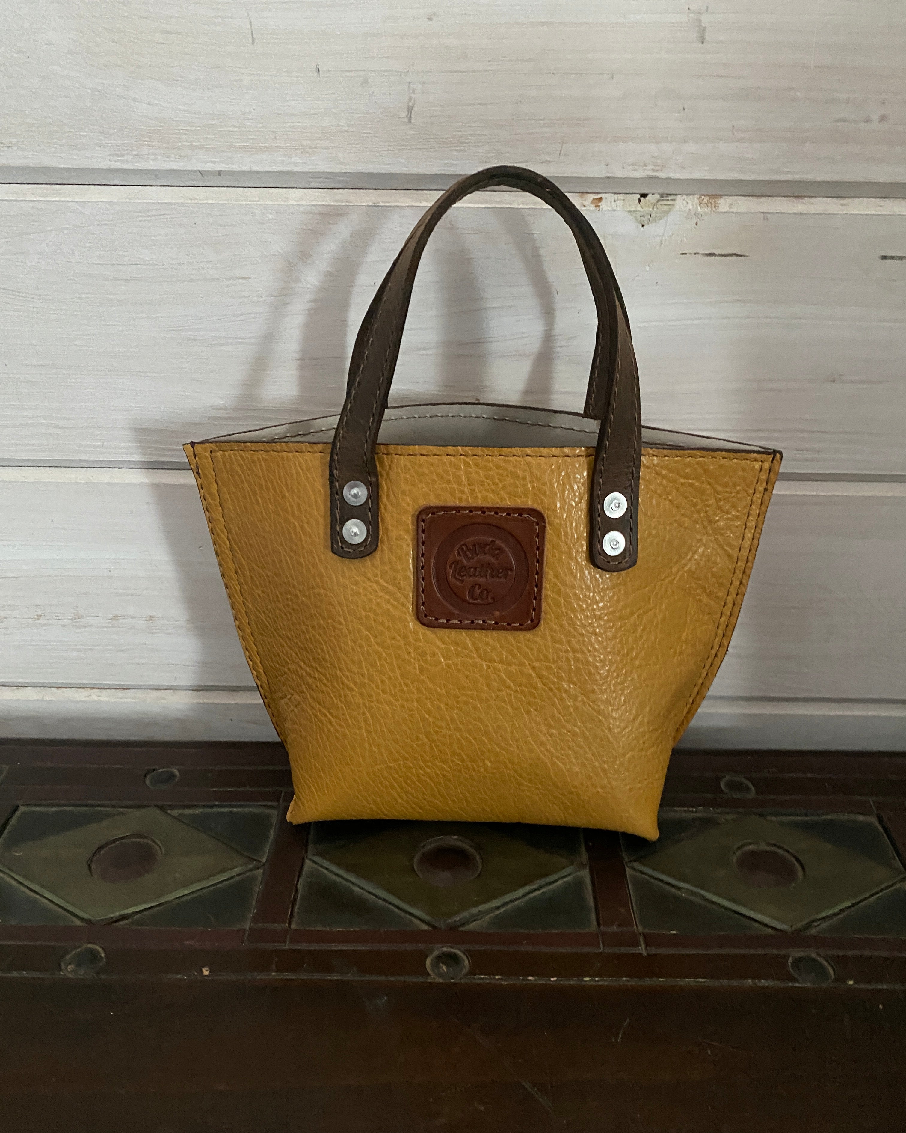 A tan pebbled leather handbag with dark brown leather handles and a brown logo patch on the front, shown against a white wood panel background.