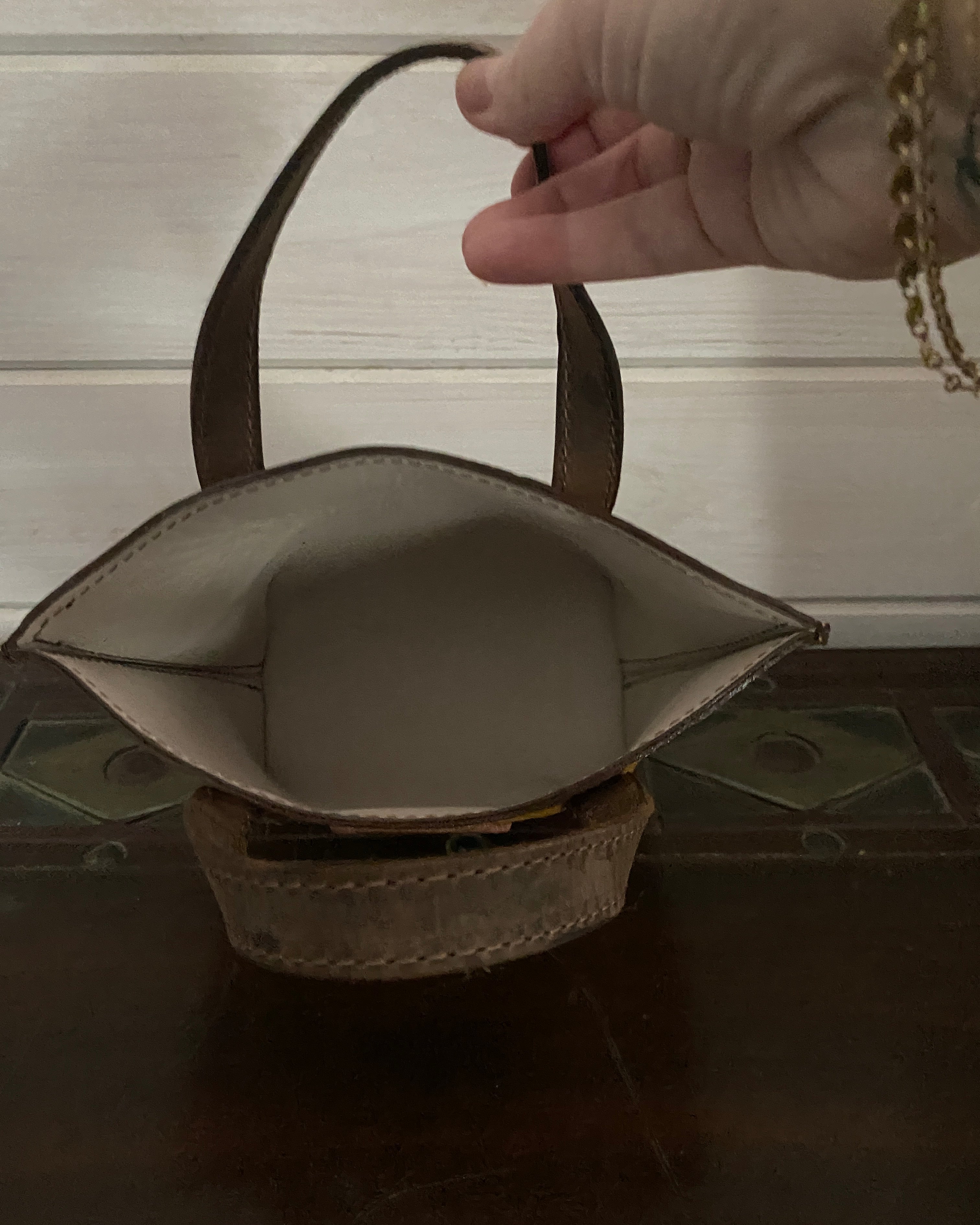 A close-up of a hand holding a small structured handbag by its strap, showing the interior lining and side profile of the bag against a dark wooden surface and light paneled background.