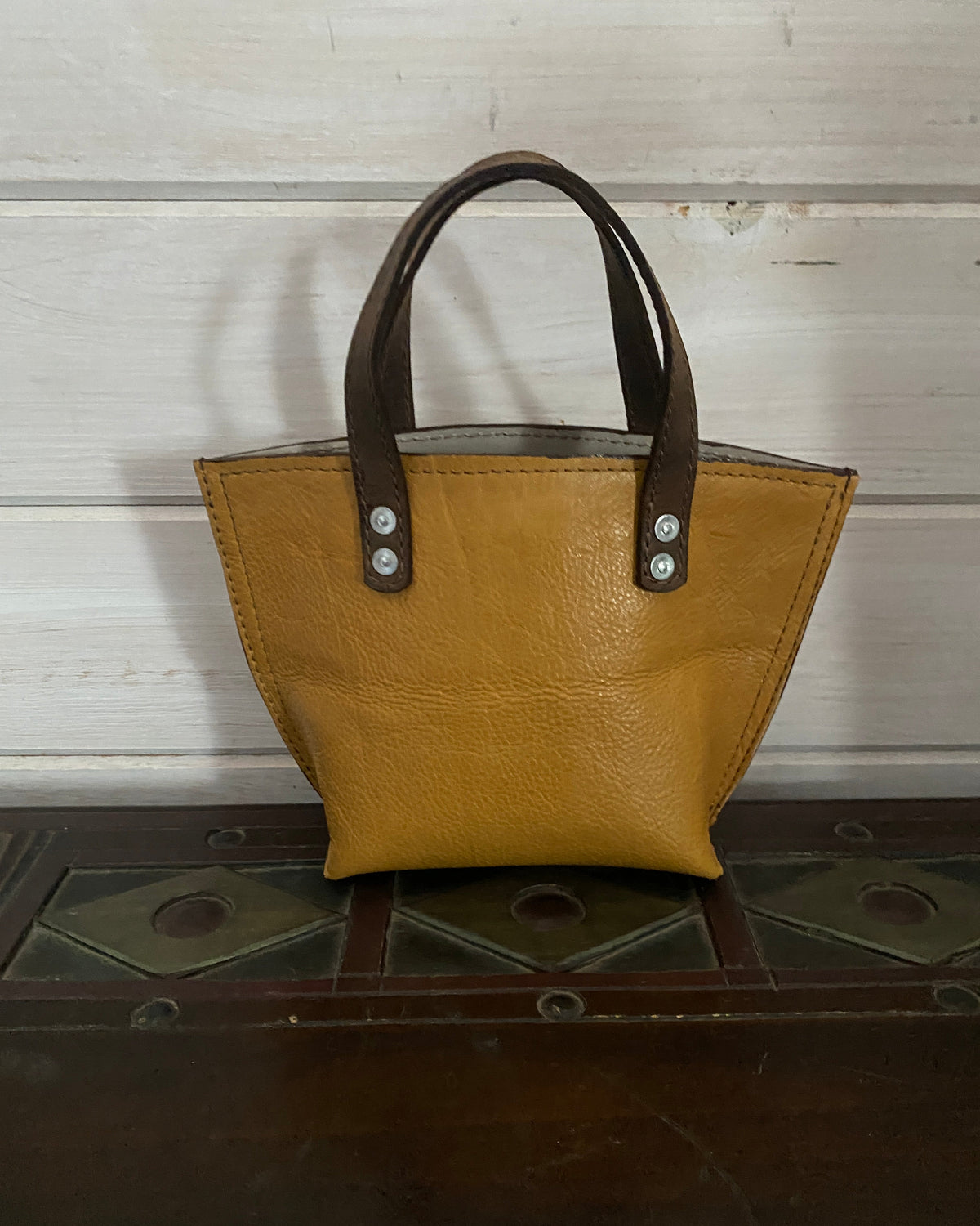 A mustard yellow leather handbag with dark brown leather handles and silver-tone hardware, shown leaning against a dark wooden surface next to a white wooden wall.