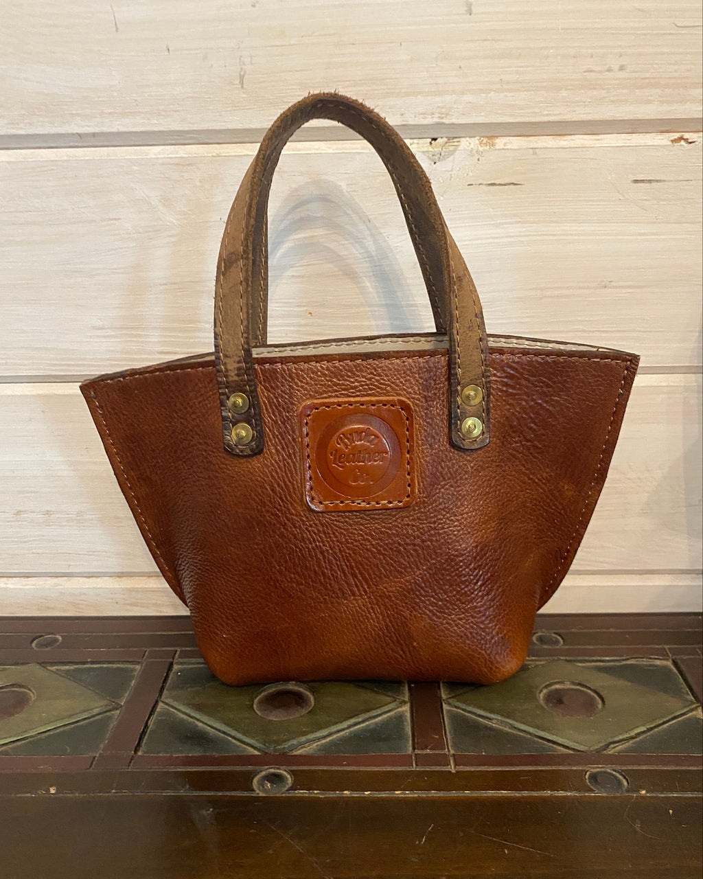 A brown leather handbag with a top handle, resting against a dark wooden surface next to a light-colored wooden panel background.