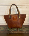 A brown leather handbag with a top handle, resting against a dark wooden surface next to a light-colored wooden panel background.