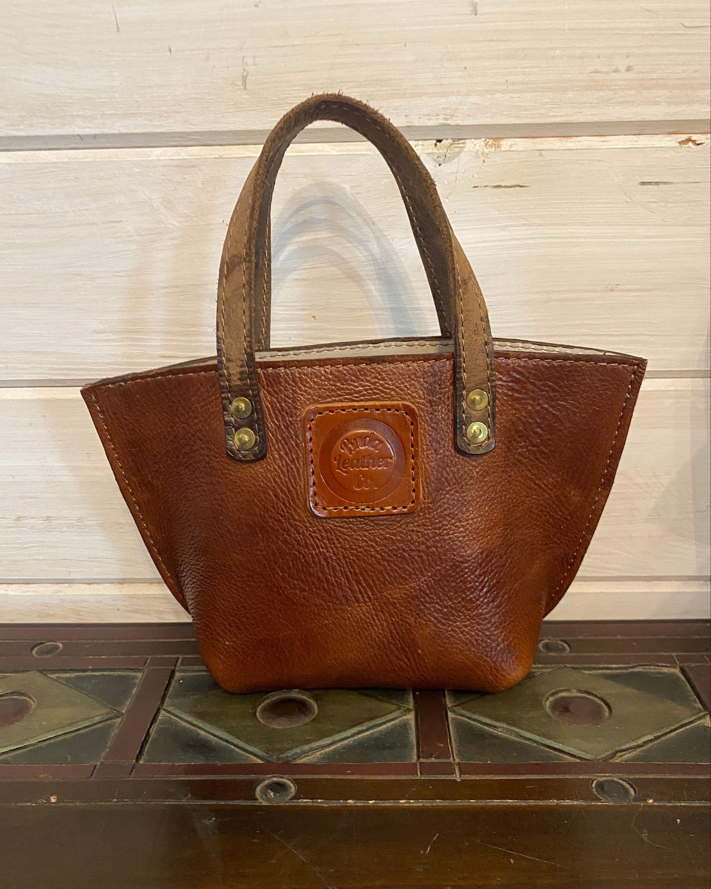 A brown leather handbag with a top handle, resting against a dark wooden surface next to a light-colored wooden panel background.