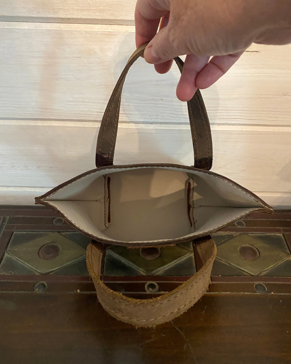 A hand holding the handle of a small brown leather tote bag, shown from a top-down view next to a dark wooden chest.