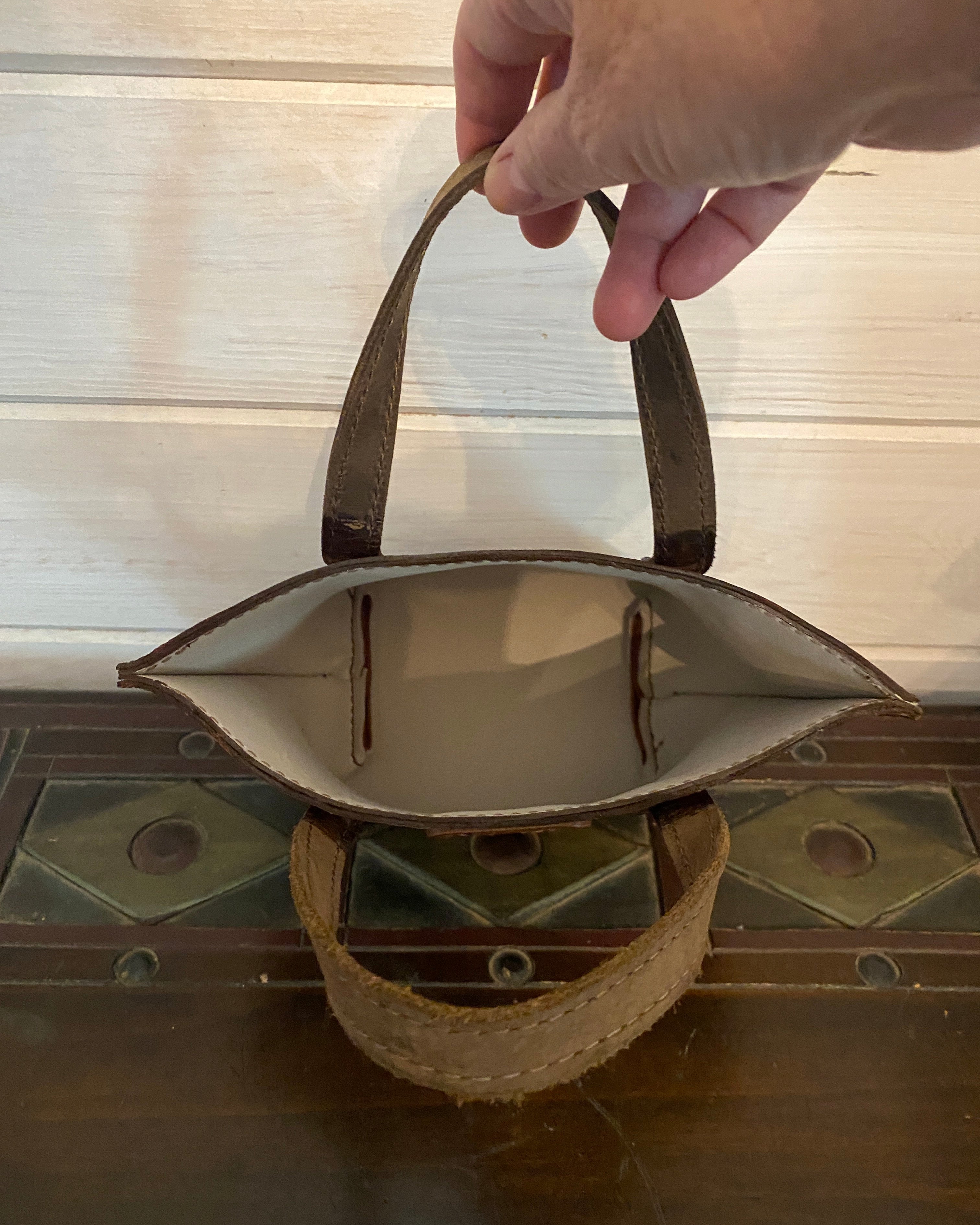 A hand holding the handle of a small brown leather tote bag, shown from a top-down view next to a dark wooden chest.