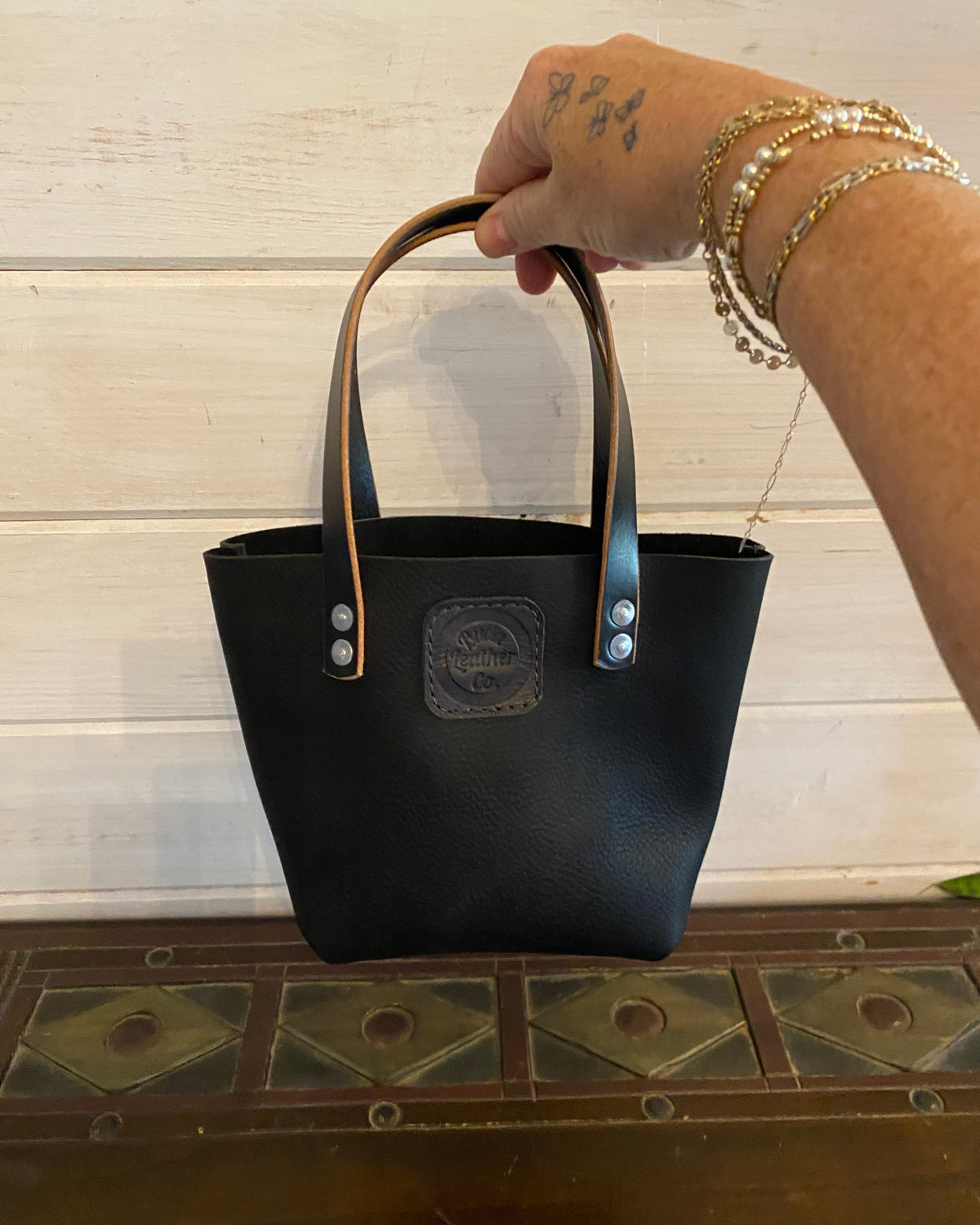 A black leather tote bag with tan handles and silver-tone hardware, held by a tattooed hand with gold and pearl bracelets against a white wood paneled wall.