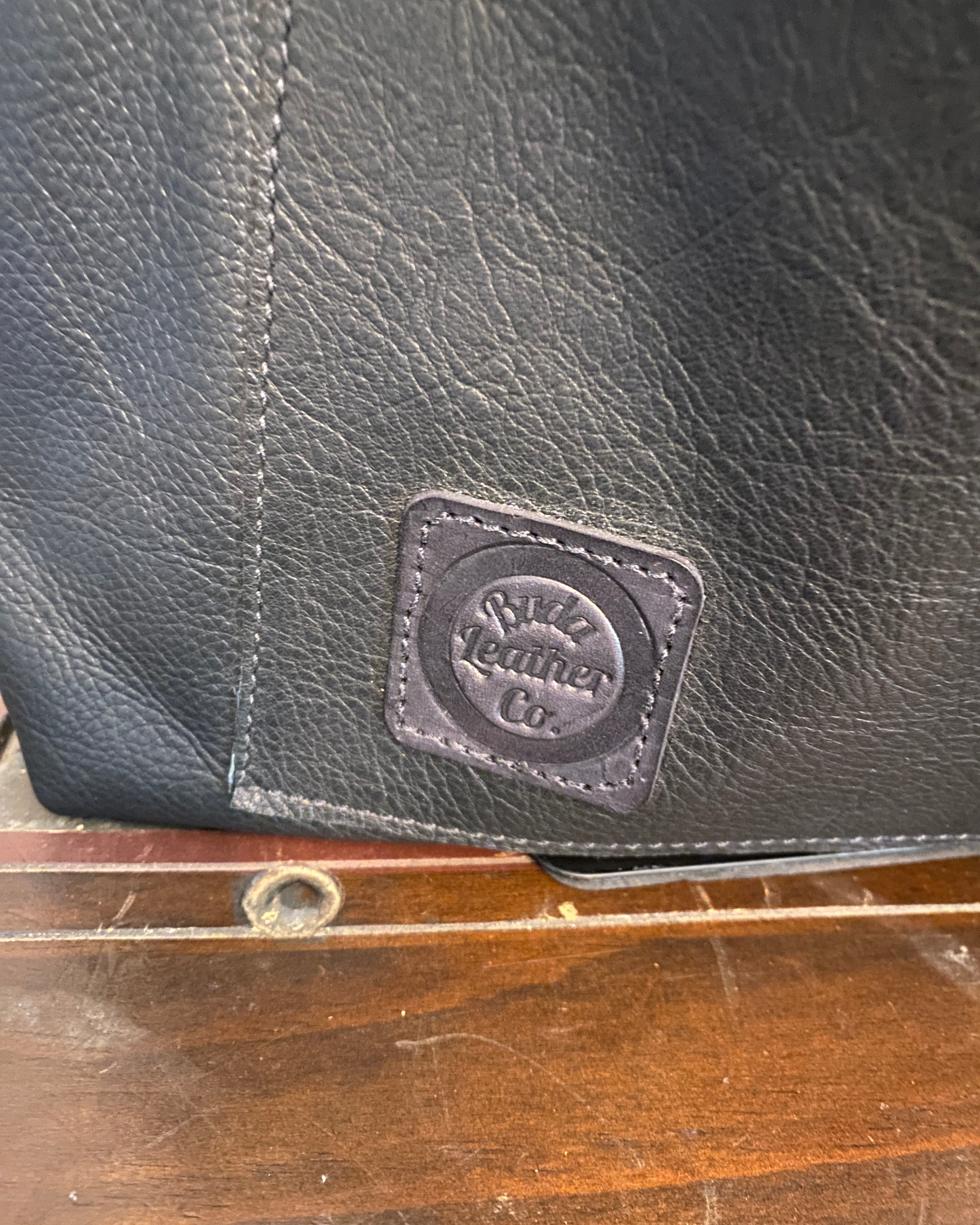 Close-up of a black leather bag with a square embossed logo patch, shown next to a wooden surface.