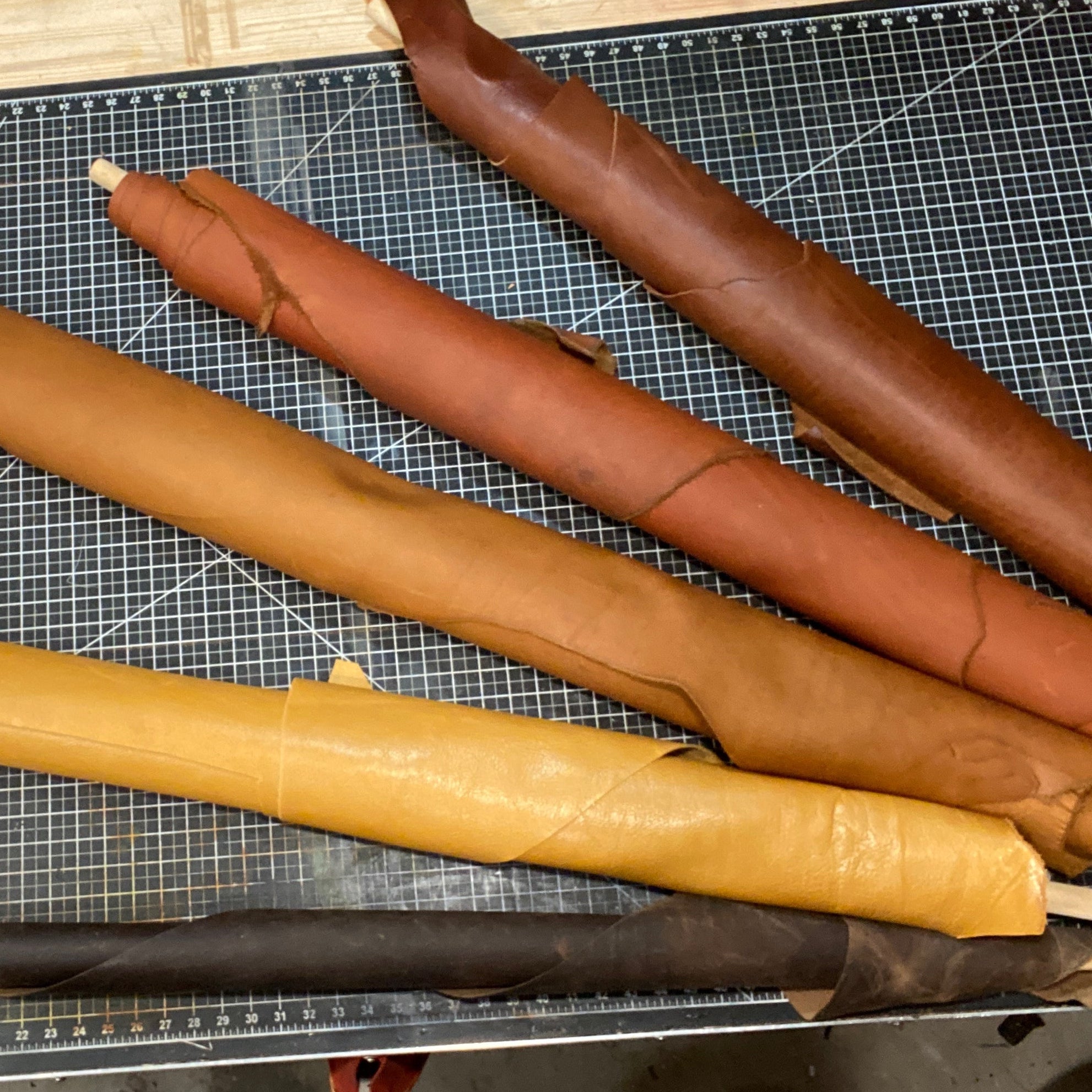 Five rolled pieces of natural leather in various shades from tan to dark brown, laid out on a black cutting mat with grid lines. A small hand tool and a piece of paper are visible near the top of the mat.