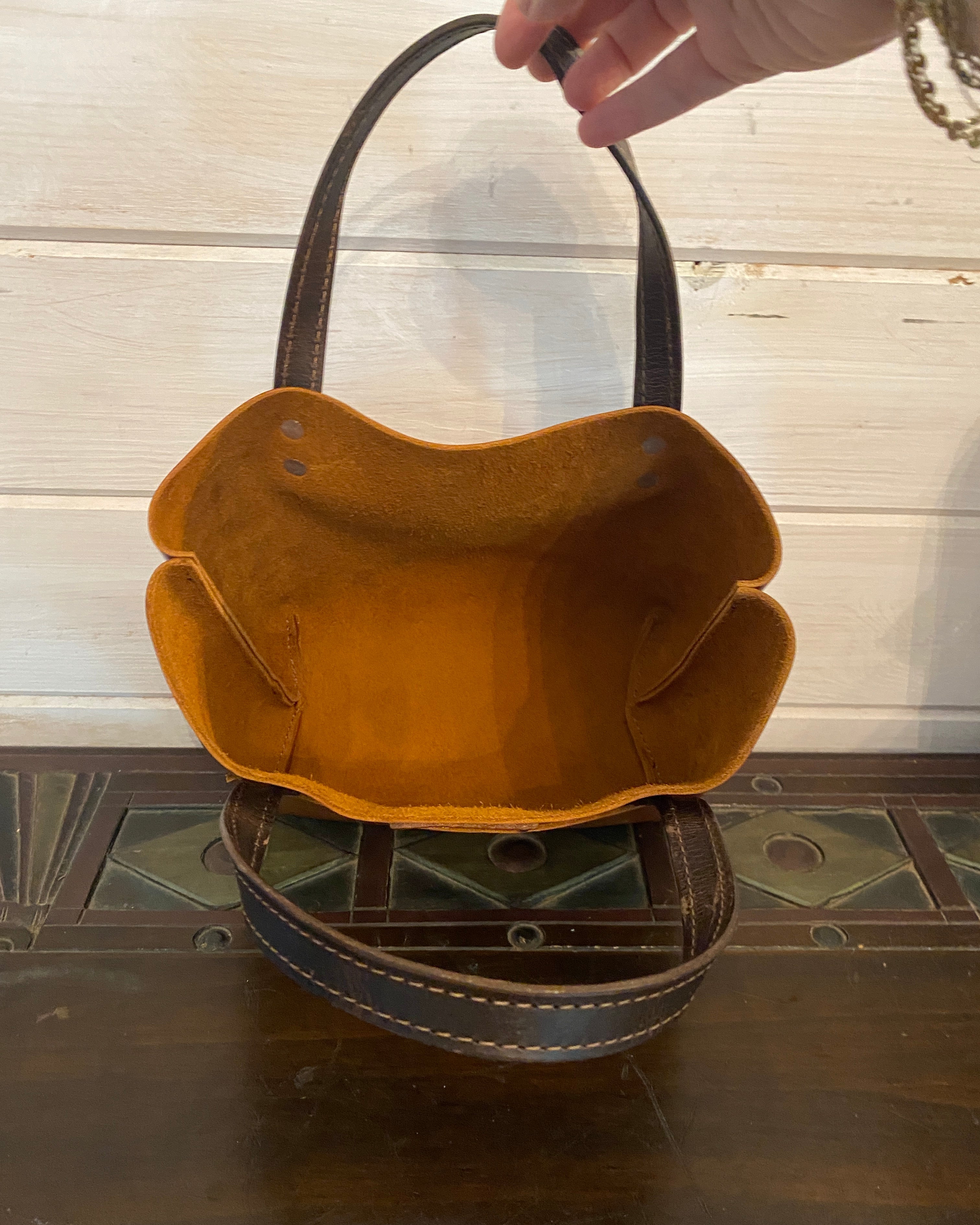 A vintage-style brown leather basket bag with a scalloped rim and dark handles, shown with a hand holding one handle against a white wood panel background.