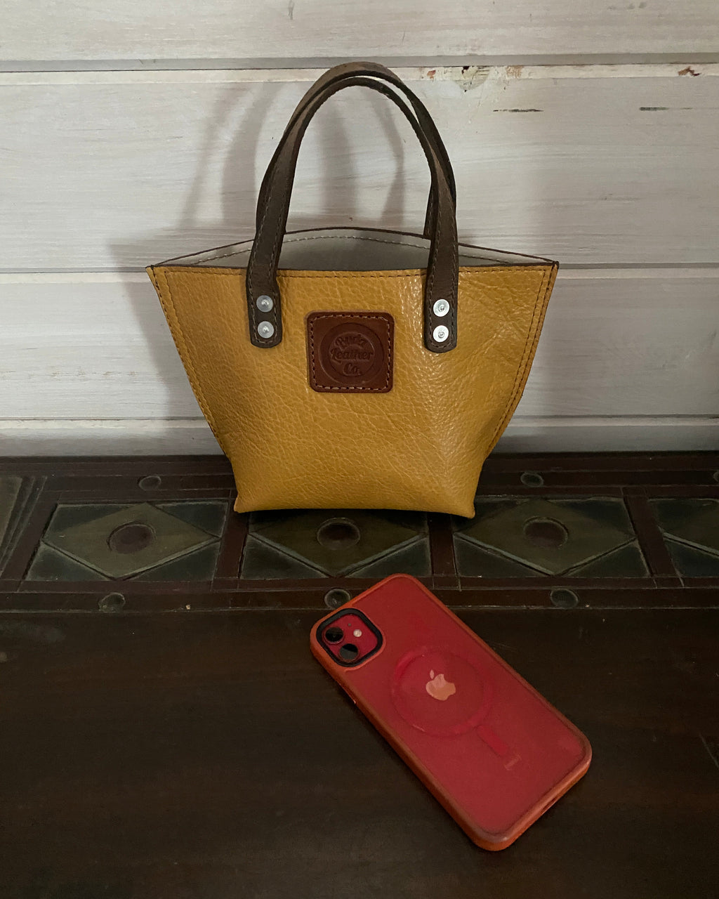 A tan pebbled leather handbag with dark brown handles and a brown logo patch on the front, shown next to a red smartphone case on a dark wooden surface.
