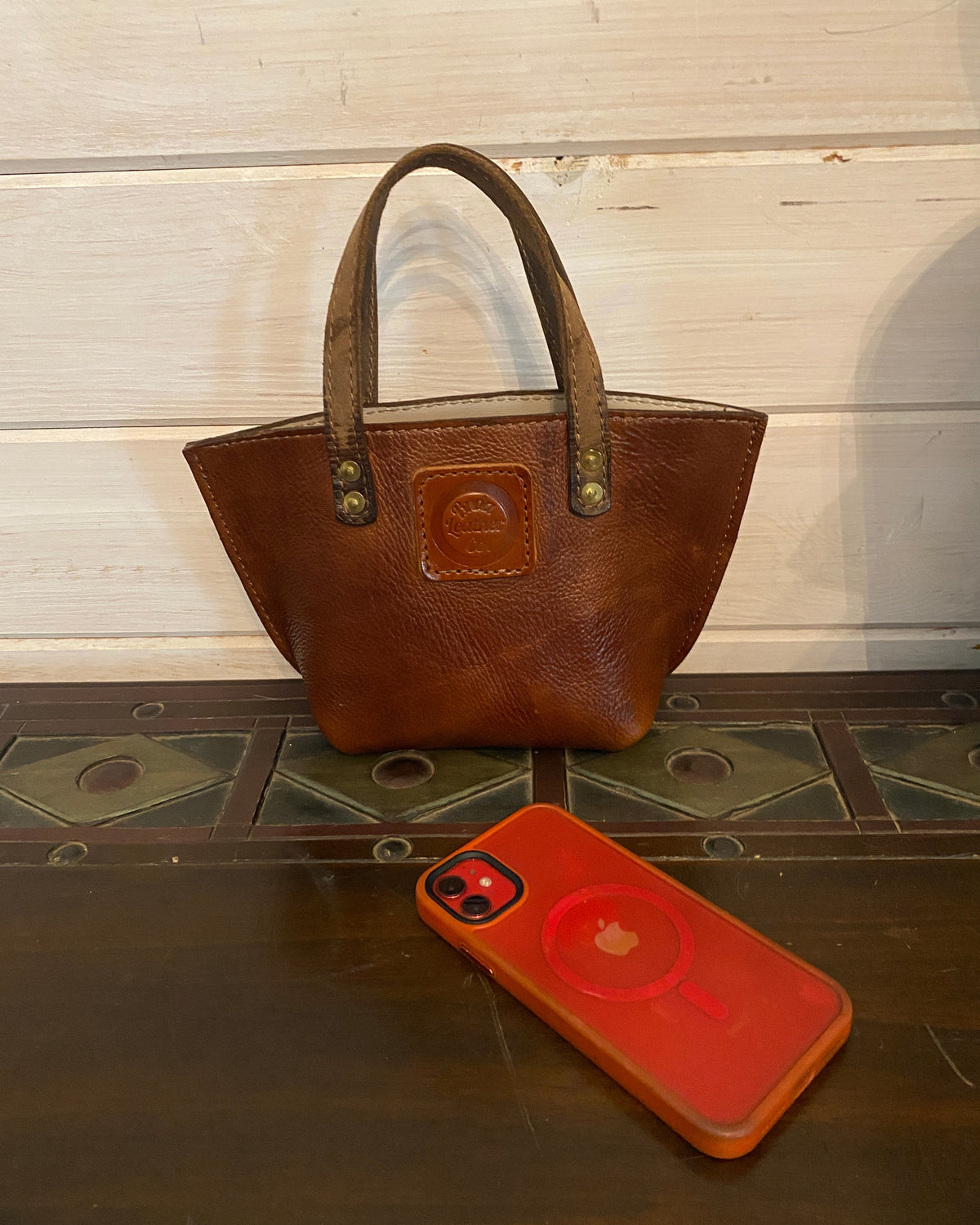 A brown leather tote bag with double handles and brass-tone hardware, shown next to a red phone case on a dark wood surface against a white paneled wall.