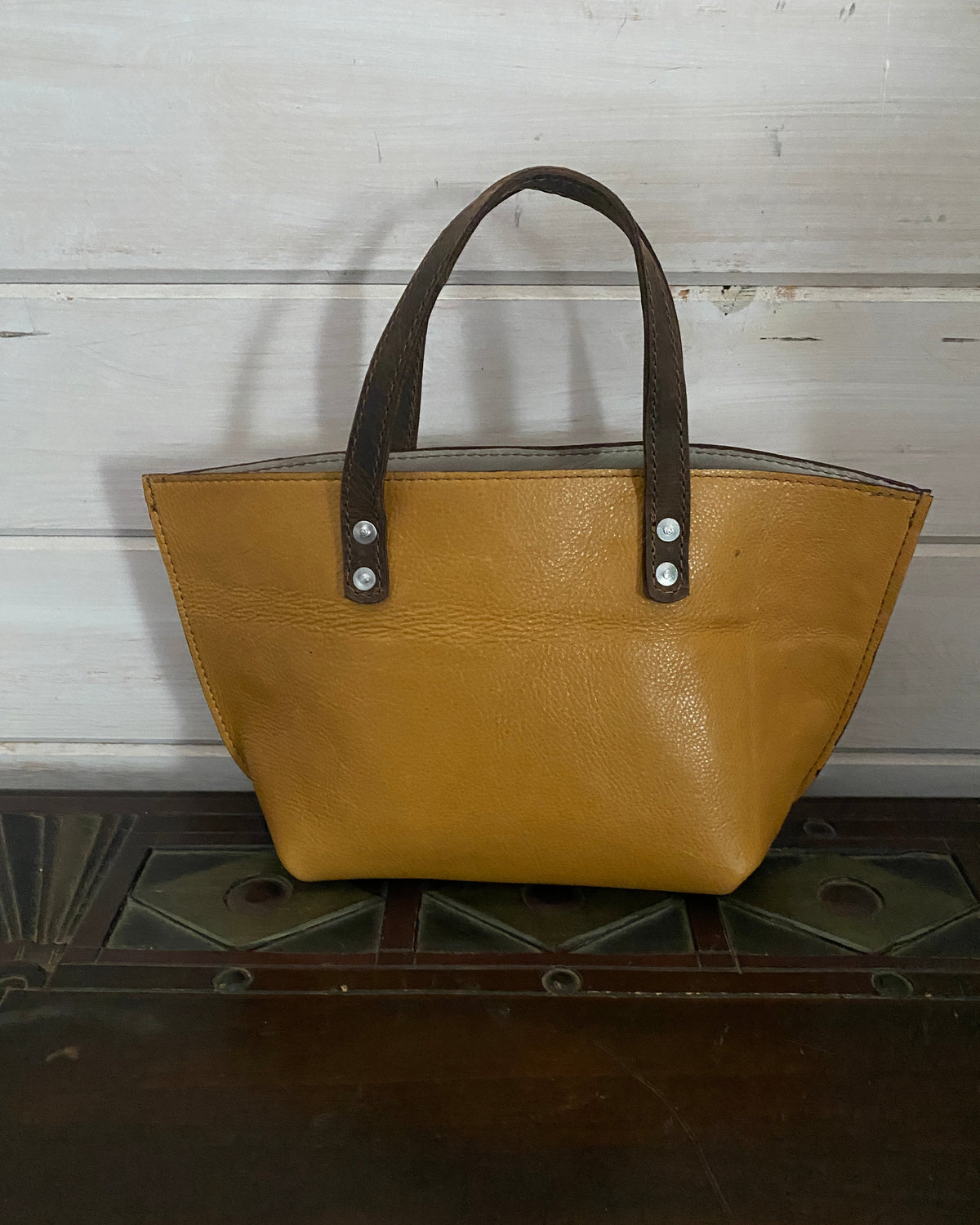 A mustard yellow leather tote bag with dark brown handles and silver-tone rivets, displayed on a dark wooden surface against a white-painted wooden wall.