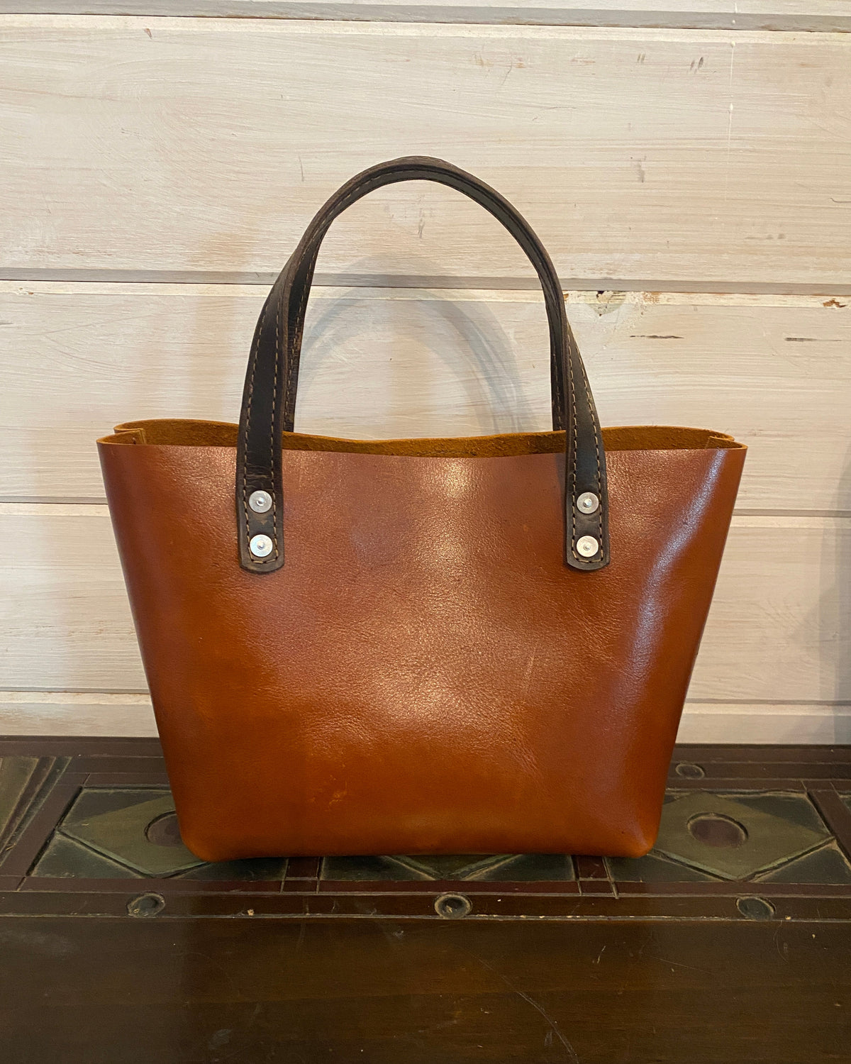 A brown leather tote bag with dark brown handles and silver-tone rivets, shown from a top-down angle on a light wood panel background next to a dark wooden surface.