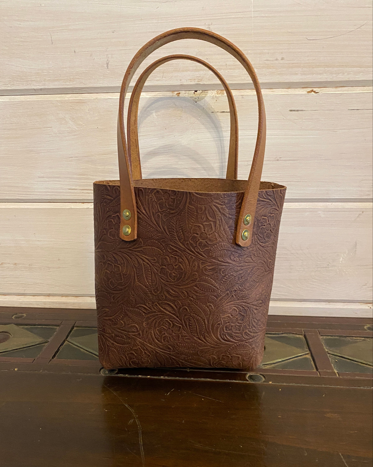 A brown embossed leather tote bag with double top handles, standing on a dark wooden table against a light wood paneled wall.