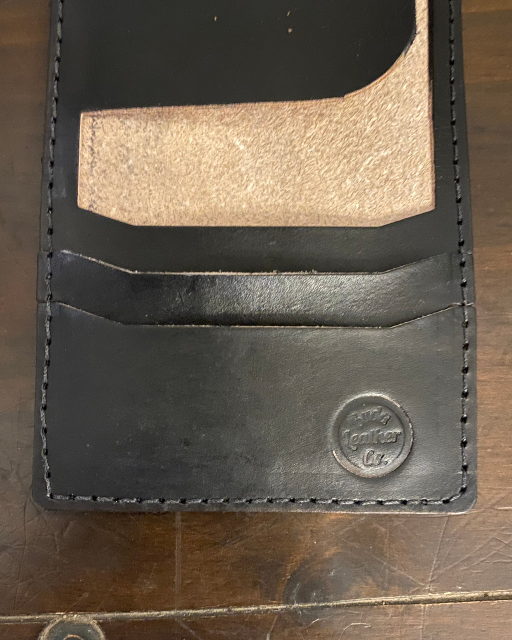 A close-up of a black leather card holder wallet with a gold-toned metallic card insert, resting on a dark wooden surface. The brand name 'Buda Leather Co.'