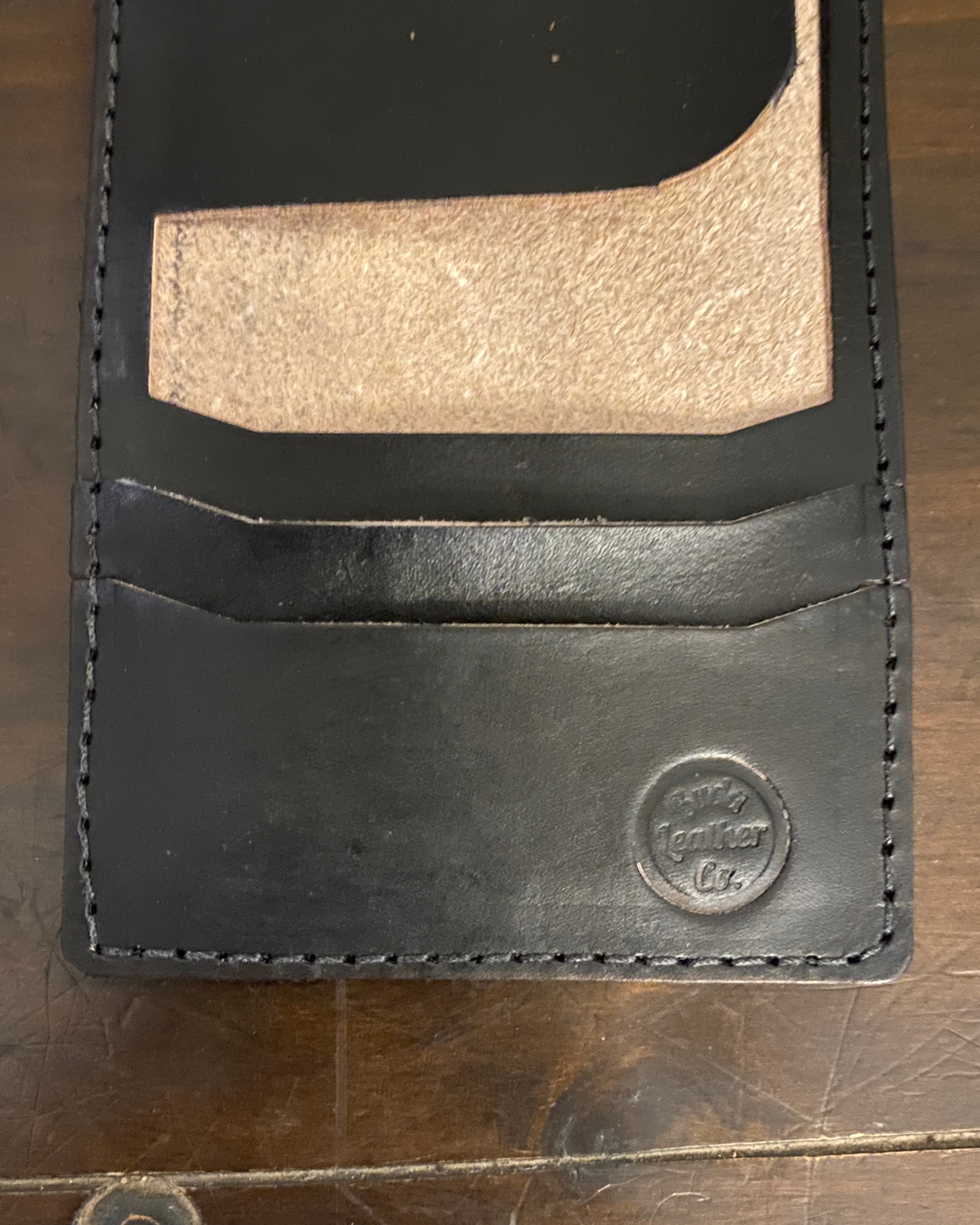 A close-up of a black leather card holder wallet with a gold-toned metallic card insert, resting on a dark wooden surface. The brand name 'Buda Leather Co.'