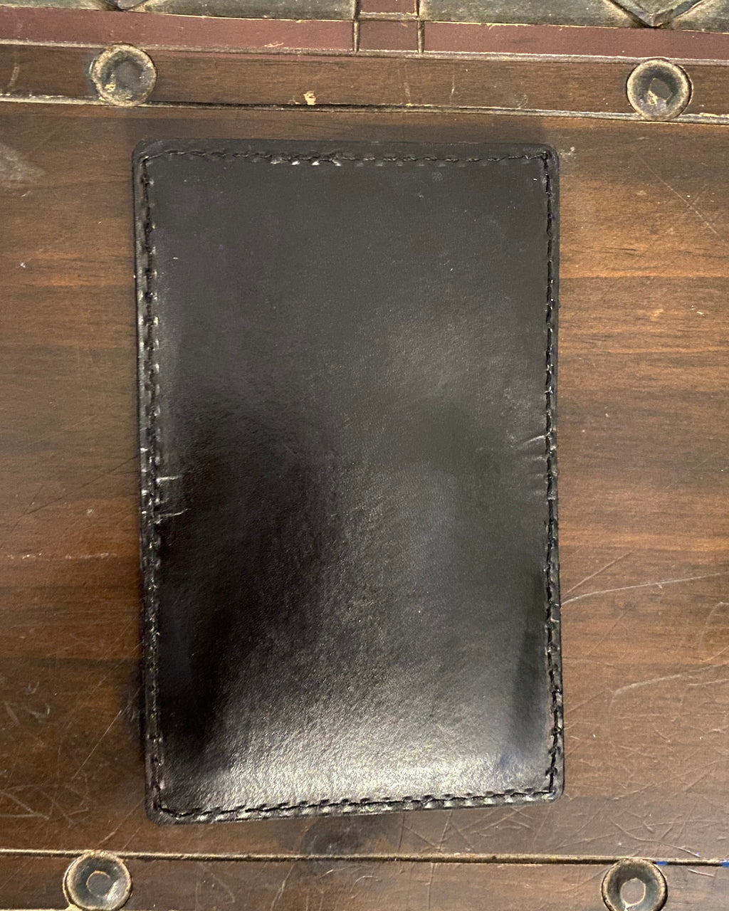 A black leather card holder or sleeve with visible stitching, placed on a rustic wooden chest.
