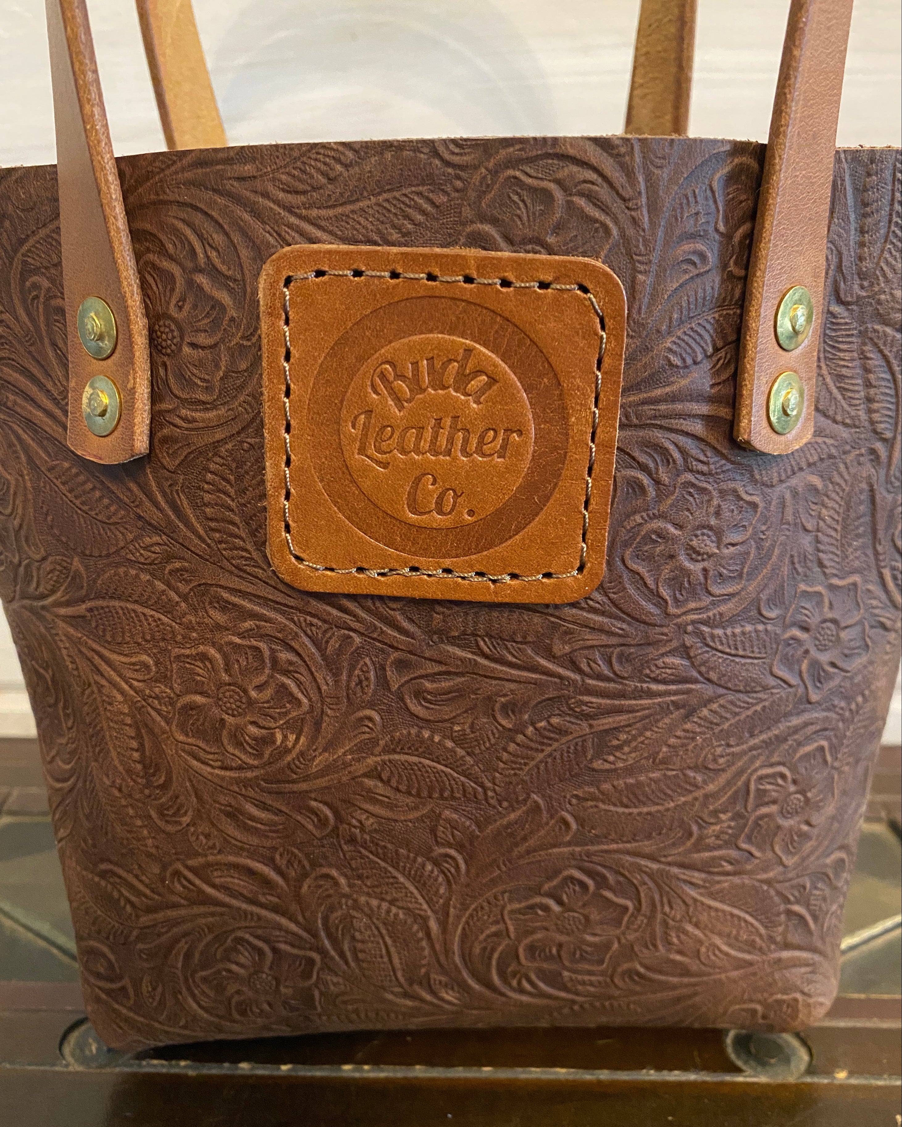 A brown tooled leather tote bag with floral embossing, featuring a tan leather patch on the front that reads 'Buda Leather Co.' and tan shoulder straps with brass-tone rivets.