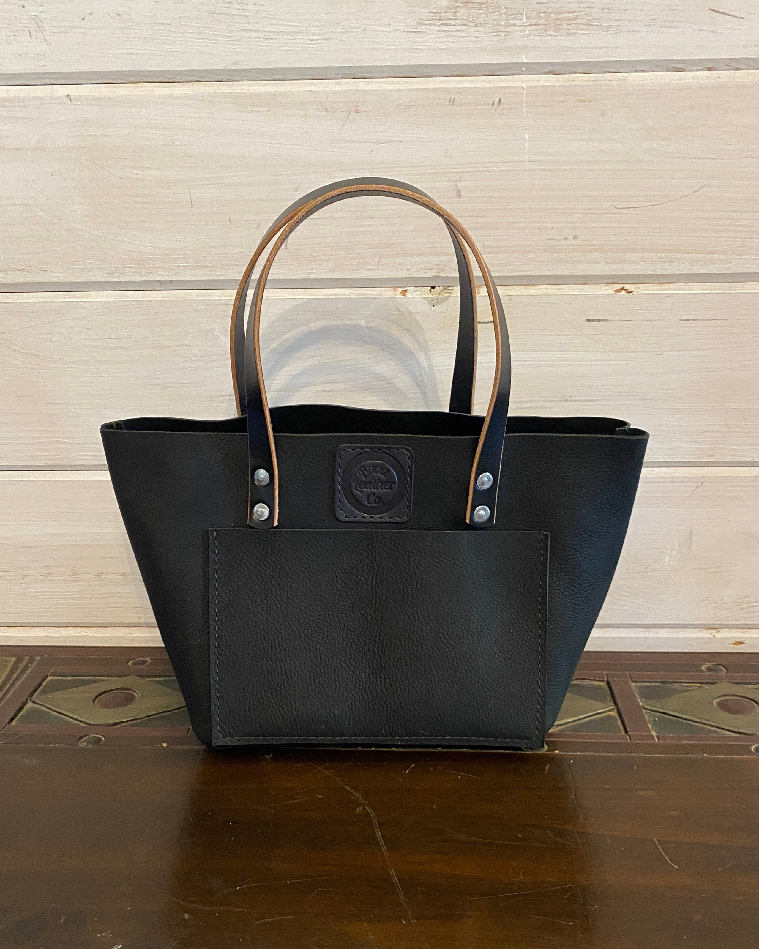 Buda Leather medium tote made from pebbled black top-grain leather with front pocket, stamped logo patch, and silver hardware.