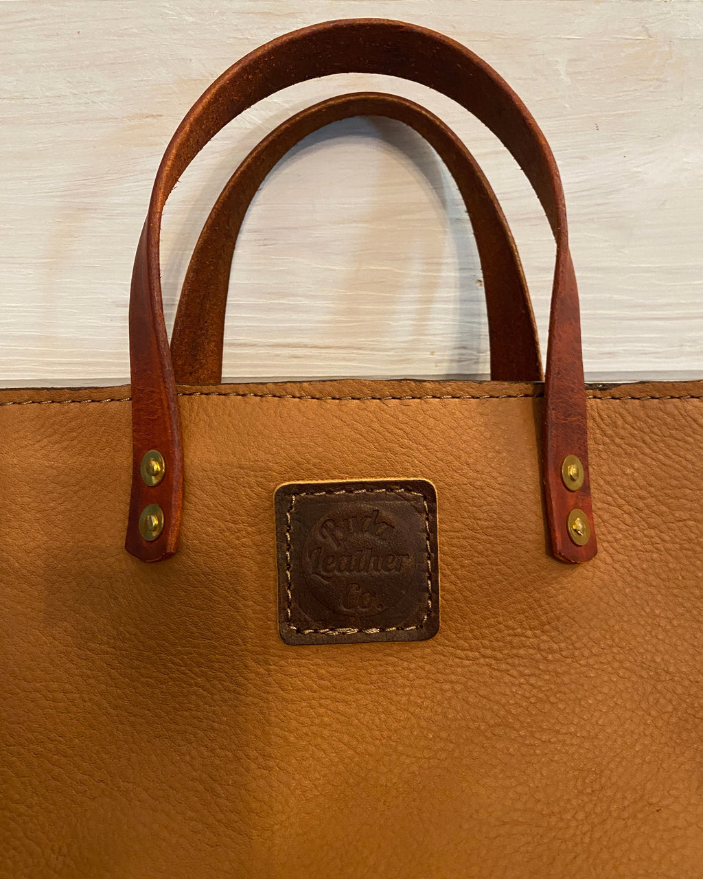 Close-up of a tan leather tote bag with dark brown leather handles and a square embossed brand patch on the front, photographed against a light wood background.