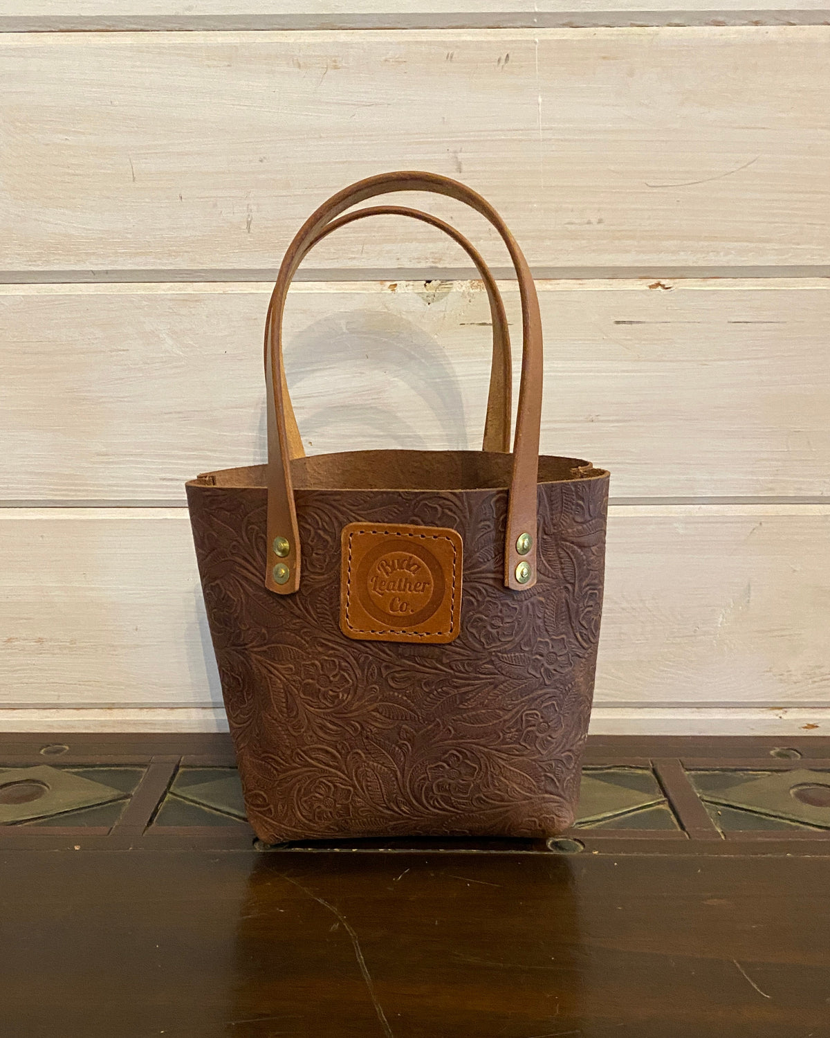 A brown embossed leather tote bag with tan handles and a tan logo patch on the front, standing on a dark wooden surface against a light wood paneled wall.