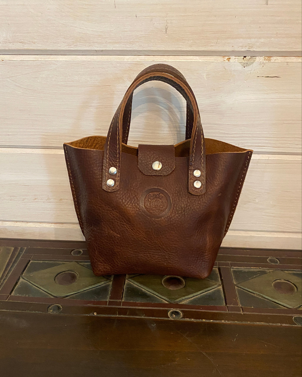 A small brown leather handbag with double top handles and silver-tone hardware, standing on a dark wooden surface against a light wood plank wall.