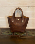 A small brown leather handbag with double top handles and silver-tone hardware, standing on a dark wooden surface against a light wood plank wall.