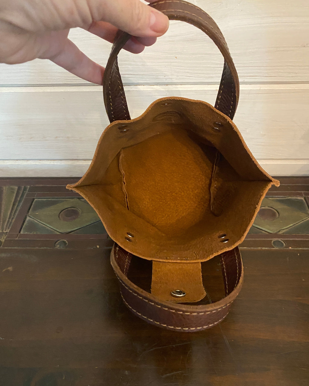 A brown leather cylindrical organizer or caddy with a hexagonal opening, held open by a hand to show the interior. The item features a dark brown exterior with visible rivets and a lighter tan interior lining, set against a dark wooden background.