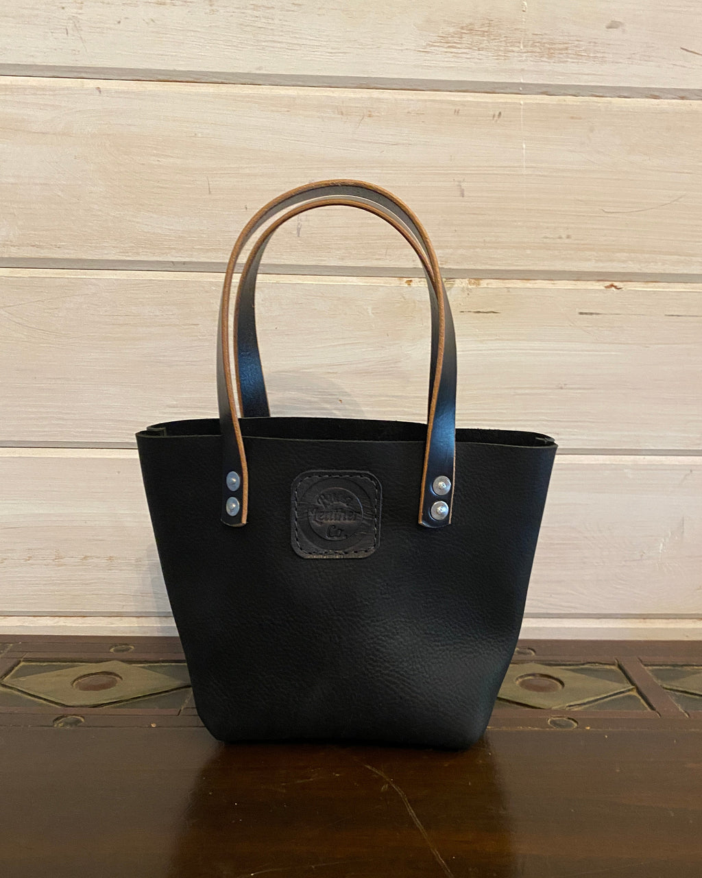 A black leather tote bag with tan handles and silver-tone hardware, standing on a dark wooden surface against a light wood plank wall.